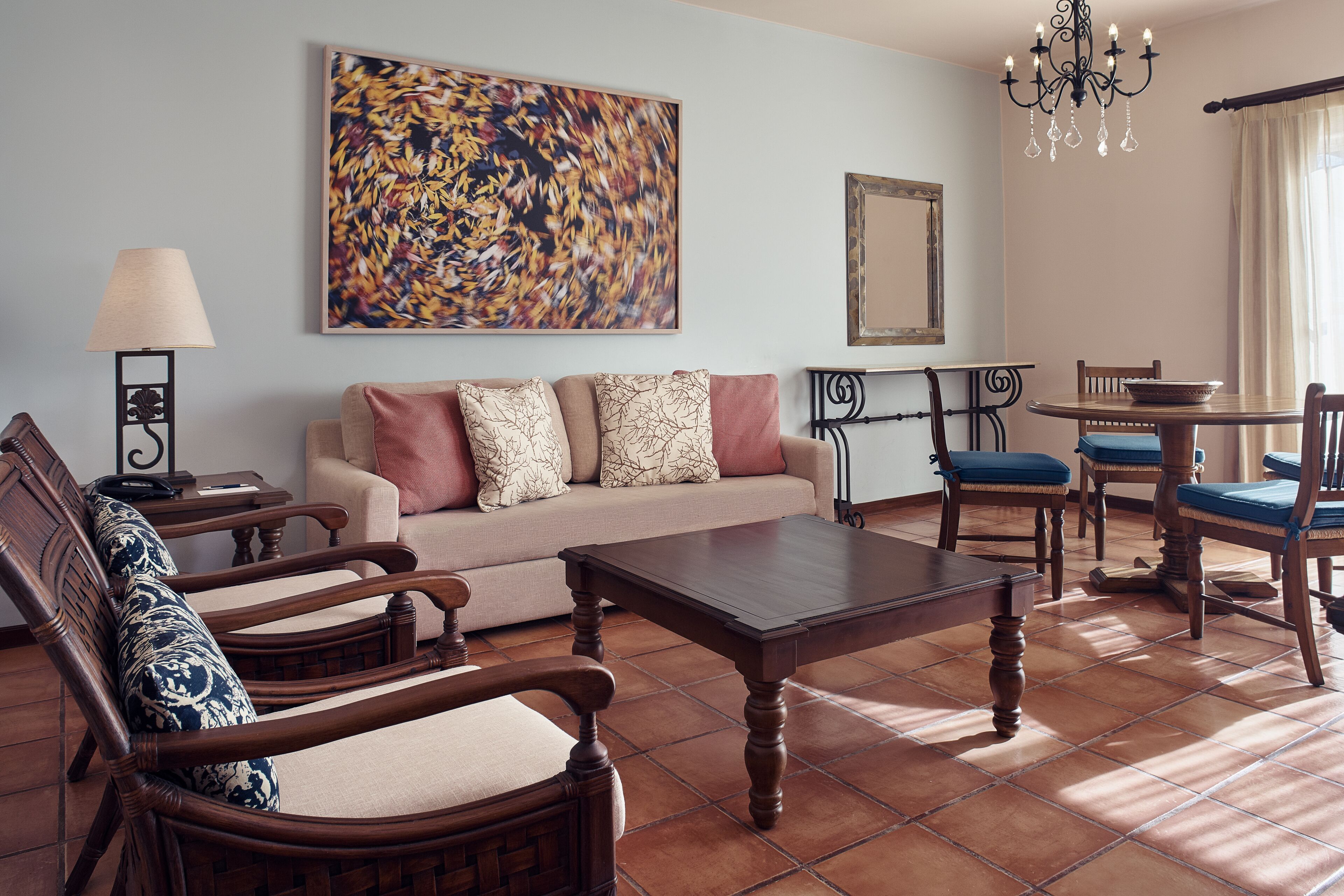Una sala de estar con sofá, dos sillones, una mesa de centro, una mesa de comedor con sillas, una araña y un cuadro abstracto grande en la pared.