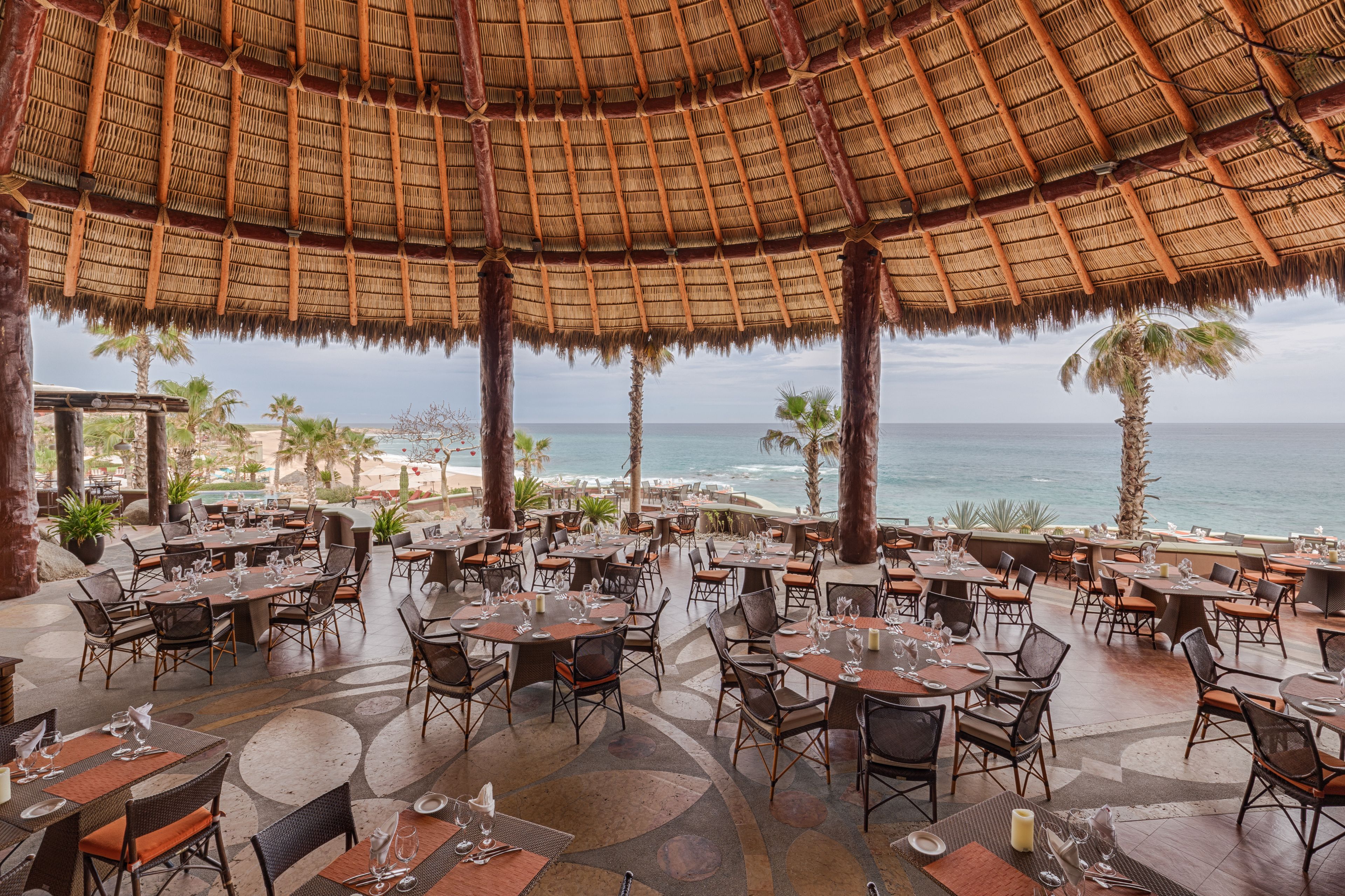 Een terras met tafels en stoelen, een rieten dak en uitzicht op de oceaan.