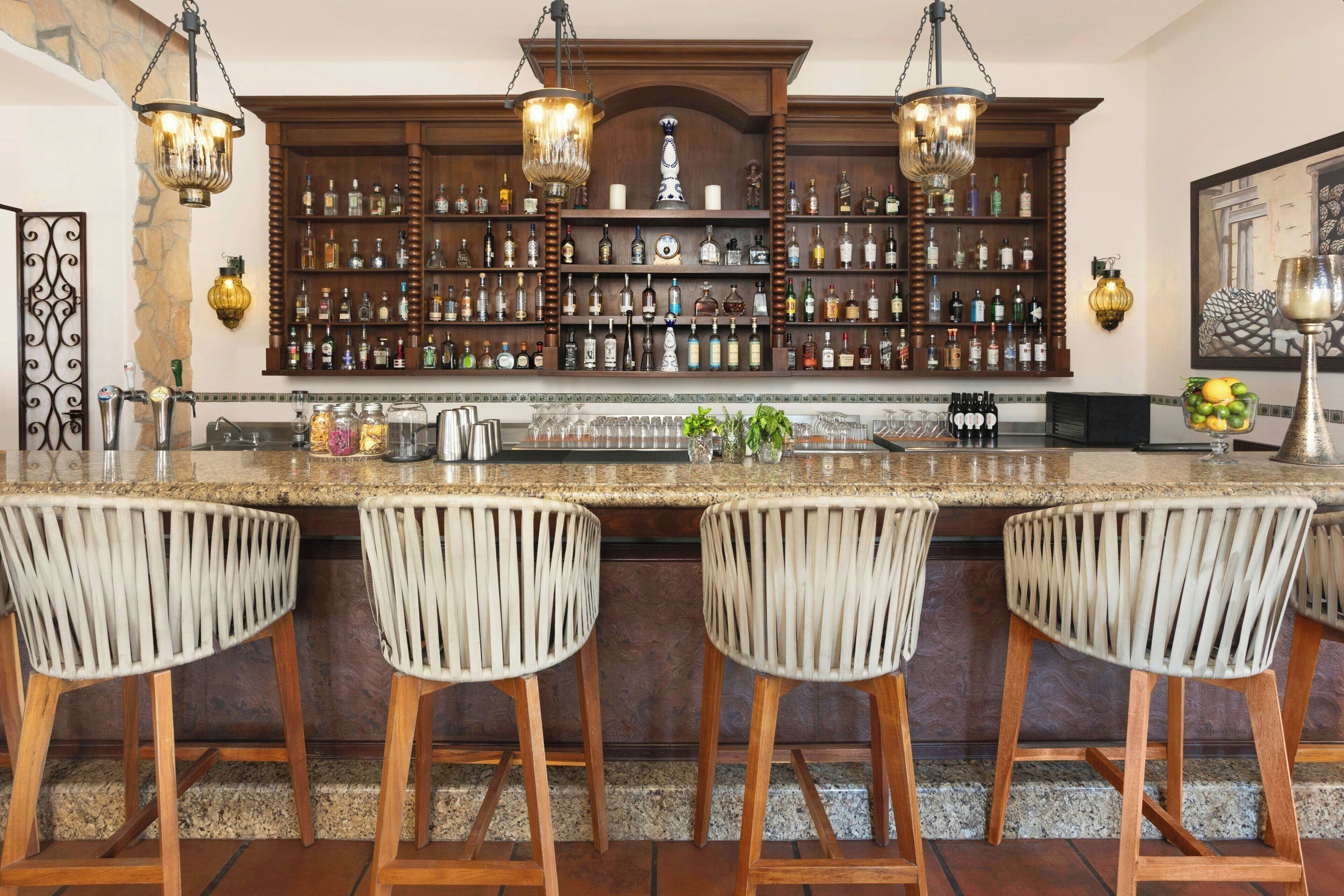 Zona de bar con encimera de granito, tres taburetes de mimbre y barra de madera con estantes llenos de botellas de licor.
