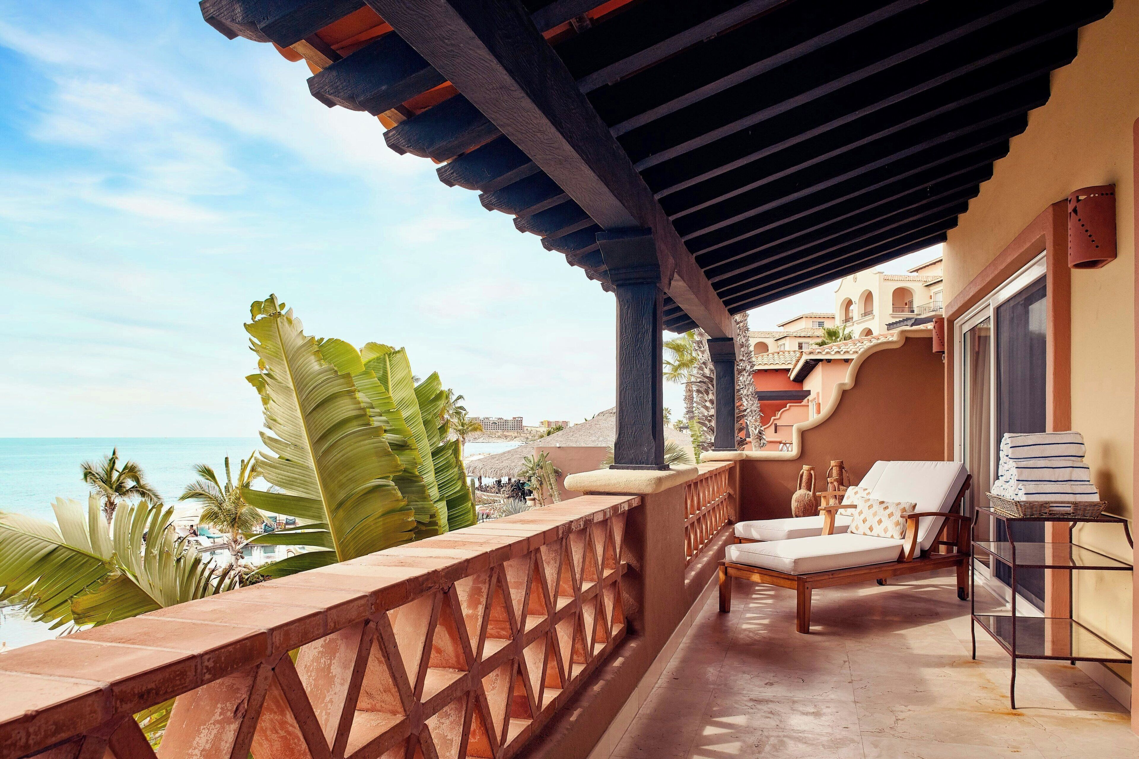 A balcony with a view of the beach, a lounge chair, and a small table with a basket.