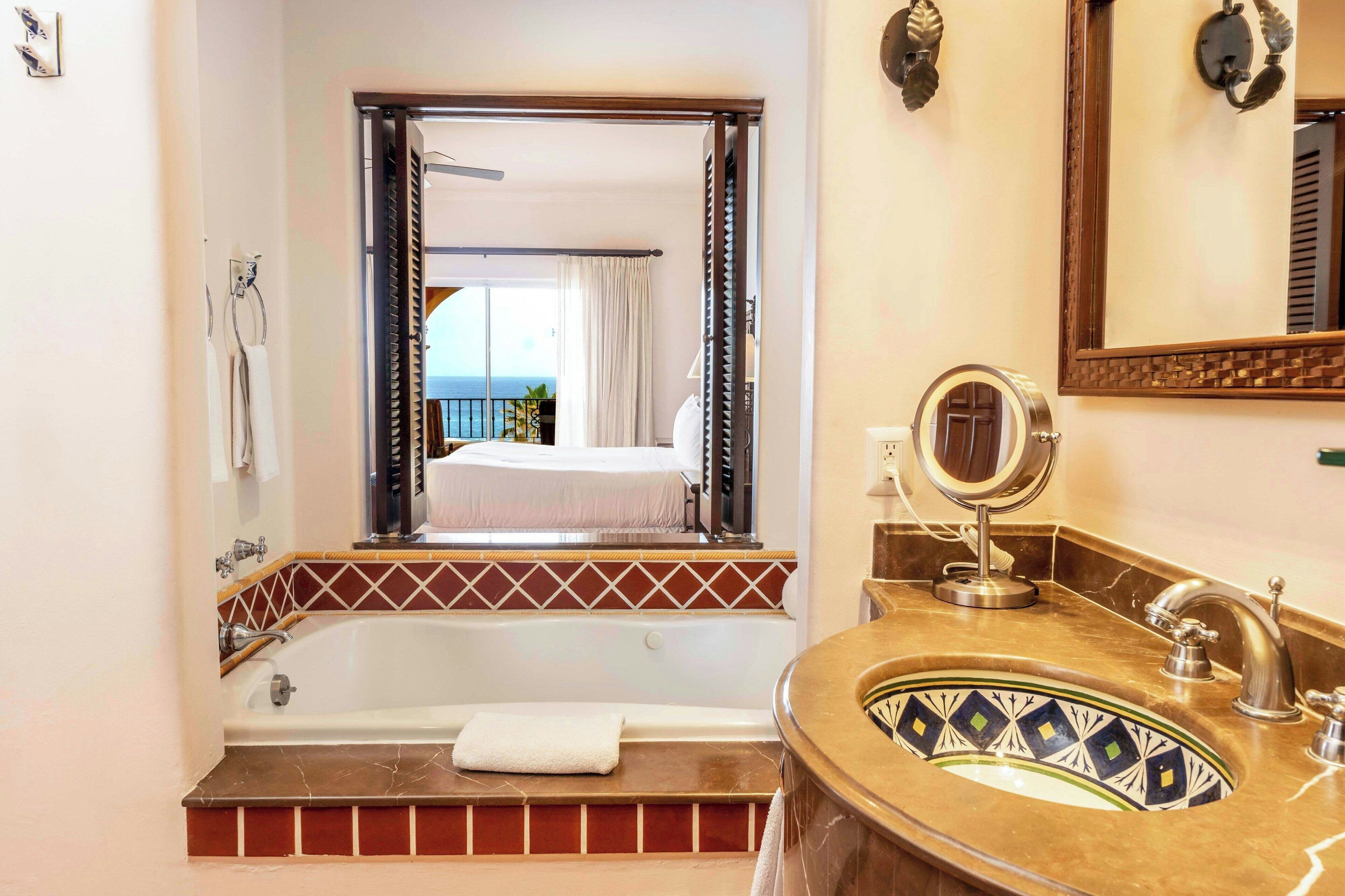 A bathroom with a bathtub, a sink with a decorative basin, a mirror, and a view of a balcony with a bed.