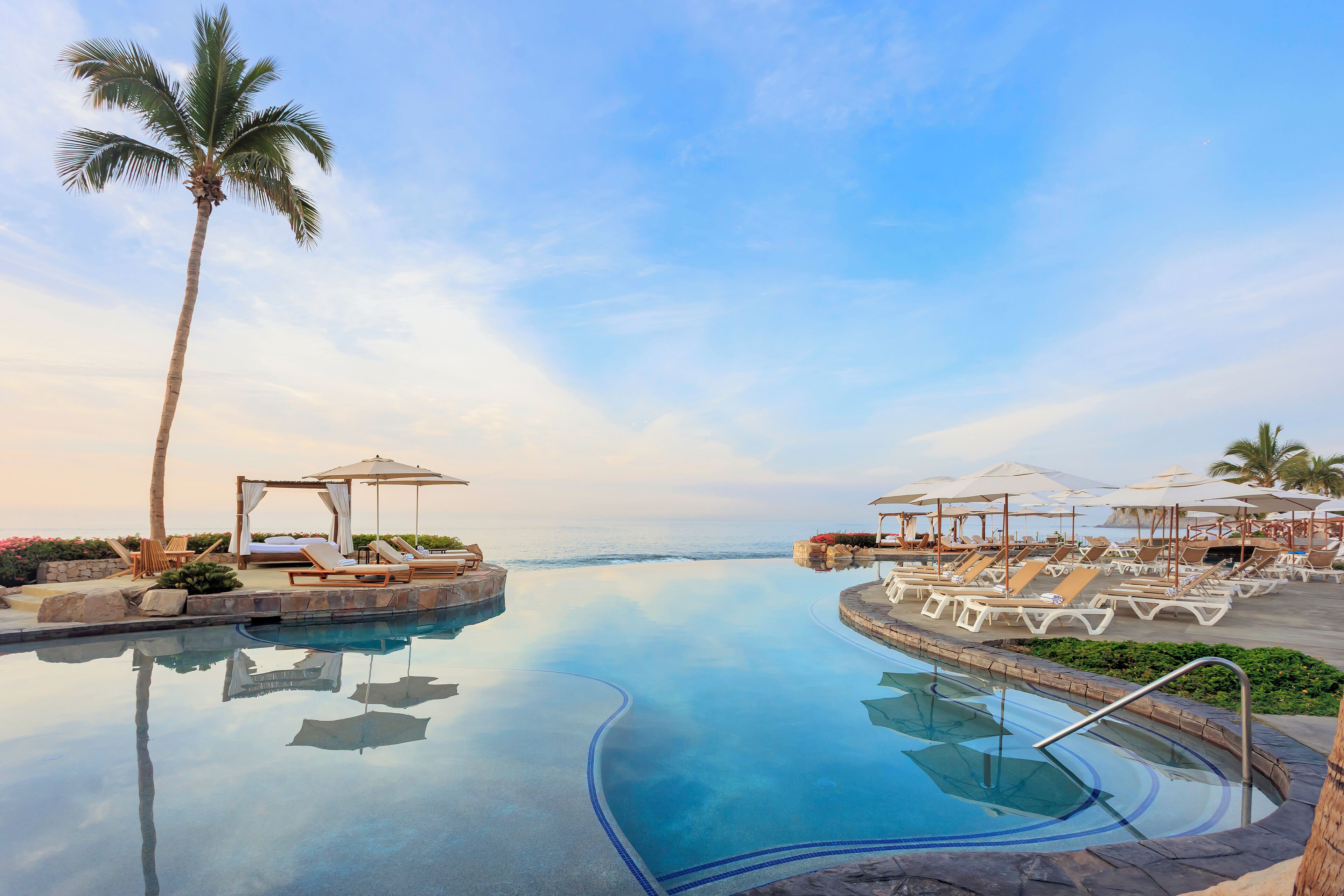 A resort pool with lounge chairs and umbrellas, palm trees, and a clear sky.
