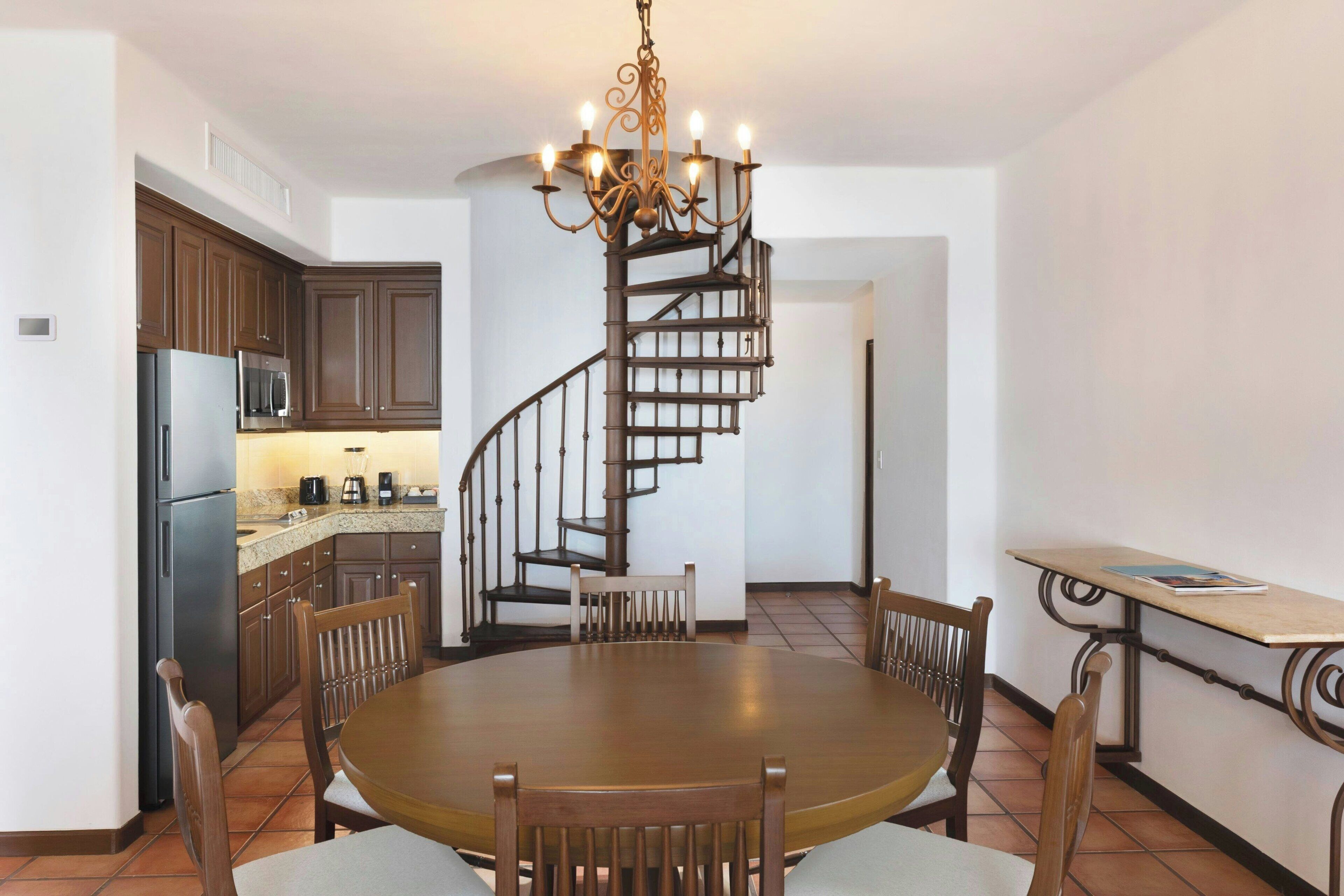 A kitchen with dark wooden cabinets, a stainless steel refrigerator, and a microwave. There is a round dining table with chairs and a chandelier above. A spiral staircase leads to an upper level.