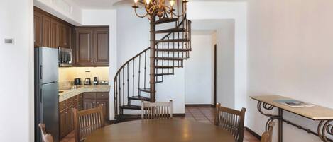 A kitchen with dark wooden cabinets, a stainless steel refrigerator, and a microwave. There is a round dining table with chairs and a chandelier above. A spiral staircase leads to an upper level.