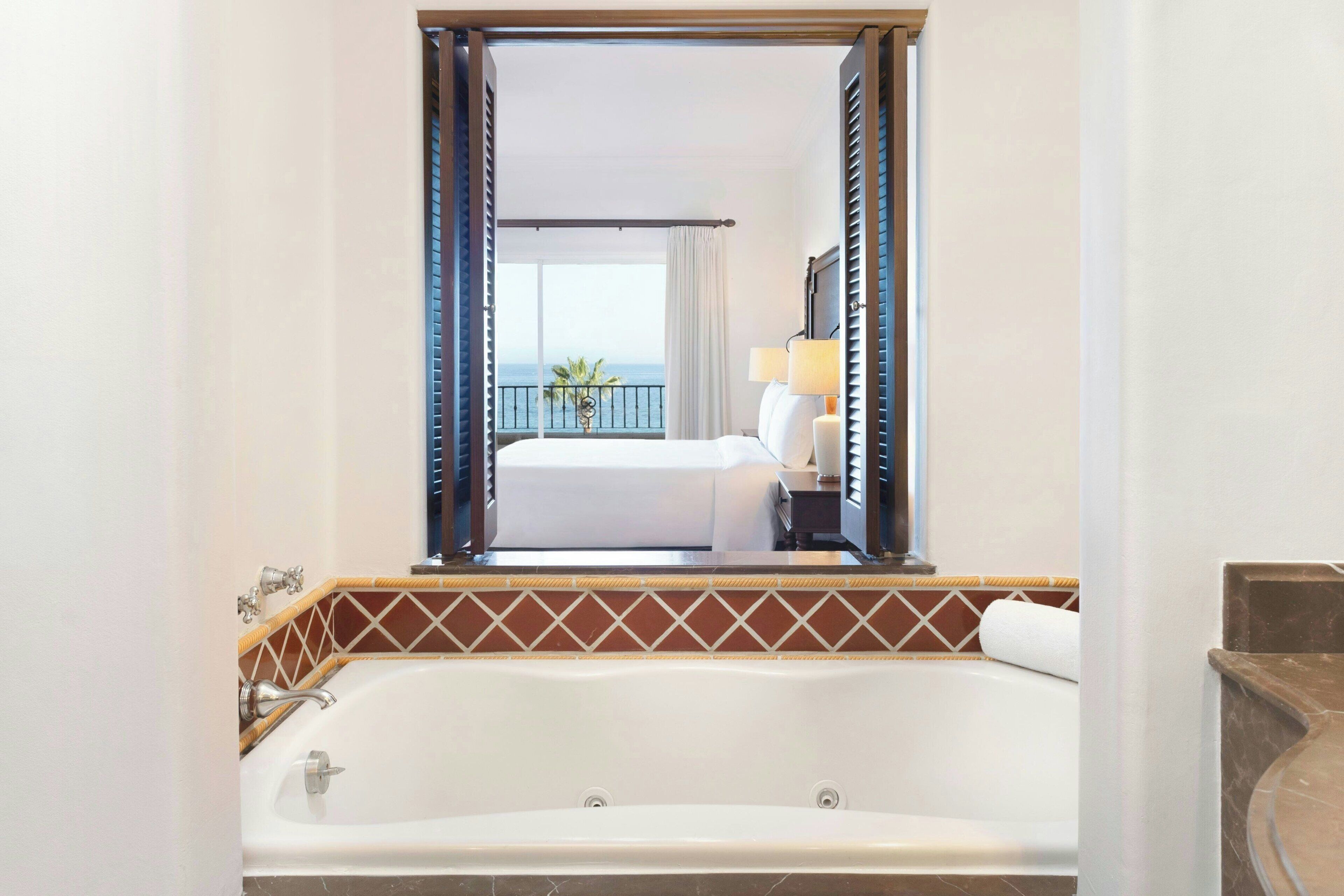 Quarto de hotel com uma banheira grande, cama com roupa de cama branca, mesa de cabeceira com abajur e varanda com vista para o mar.
