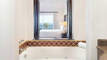 Quarto de hotel com uma banheira grande, cama com roupa de cama branca, mesa de cabeceira com abajur e varanda com vista para o mar.
