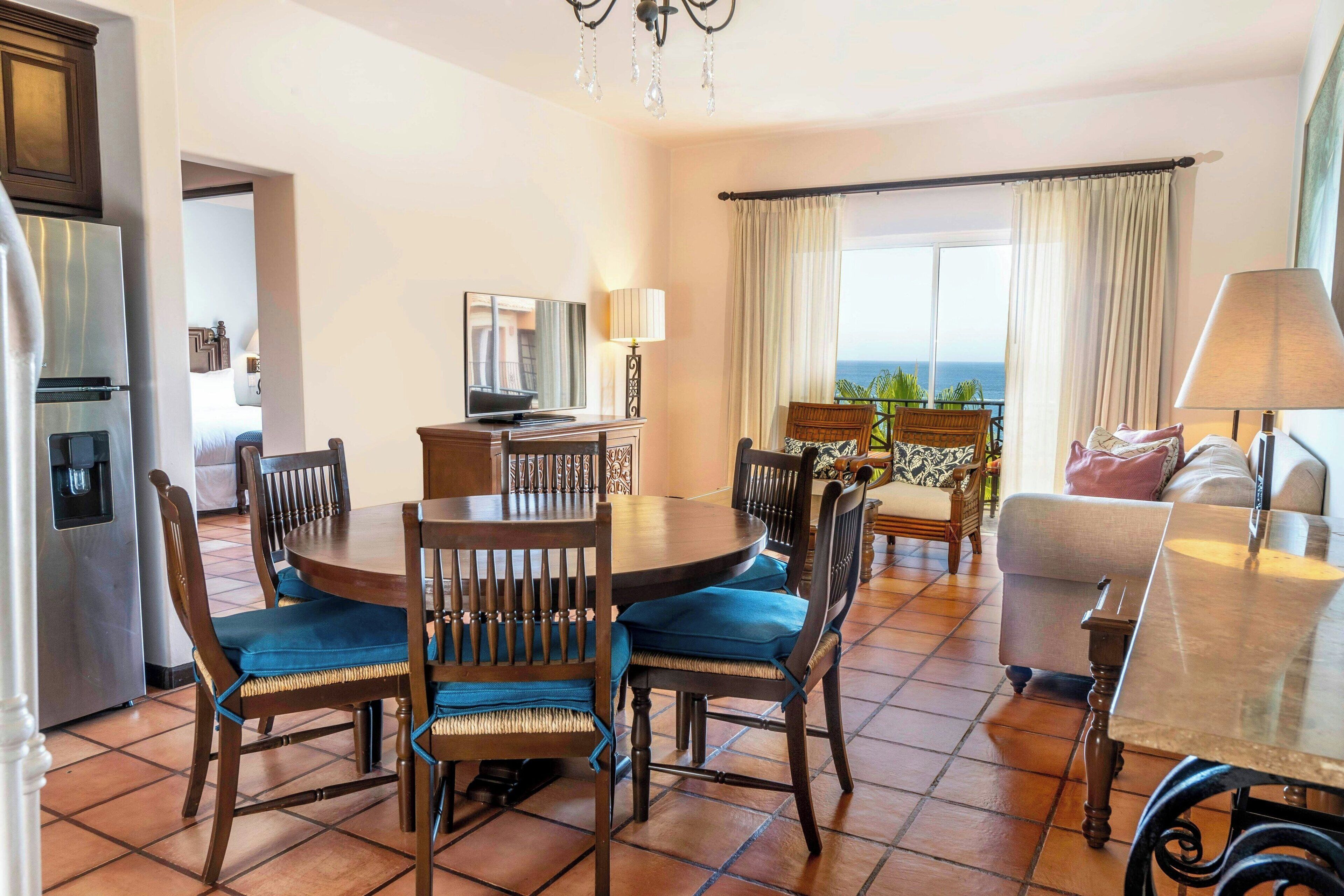 A dining area with a round table and chairs, a television, and a view of the ocean.