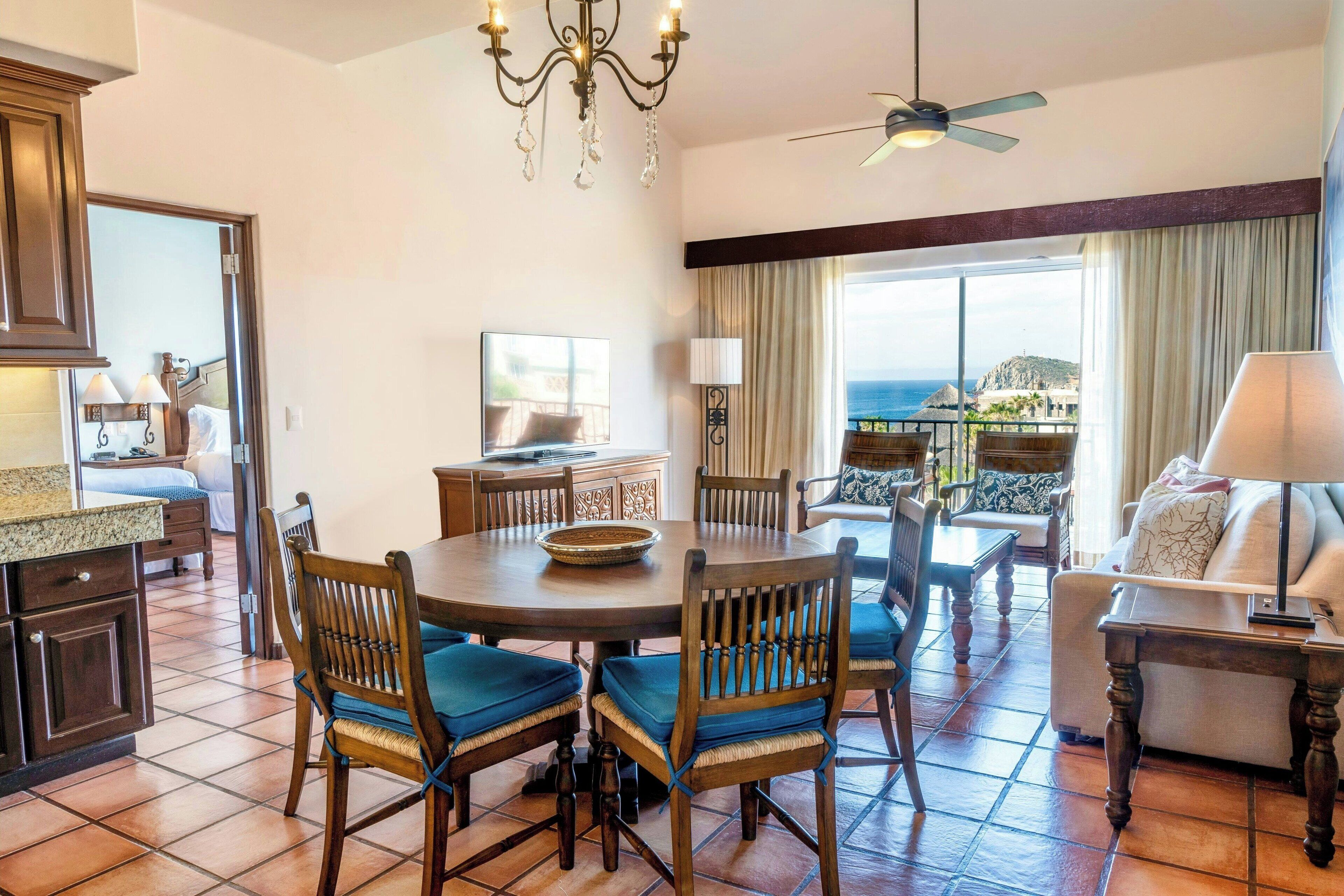 A dining area with a round table, chairs, and a chandelier. There is a television on the wall and a view of the ocean through large windows.