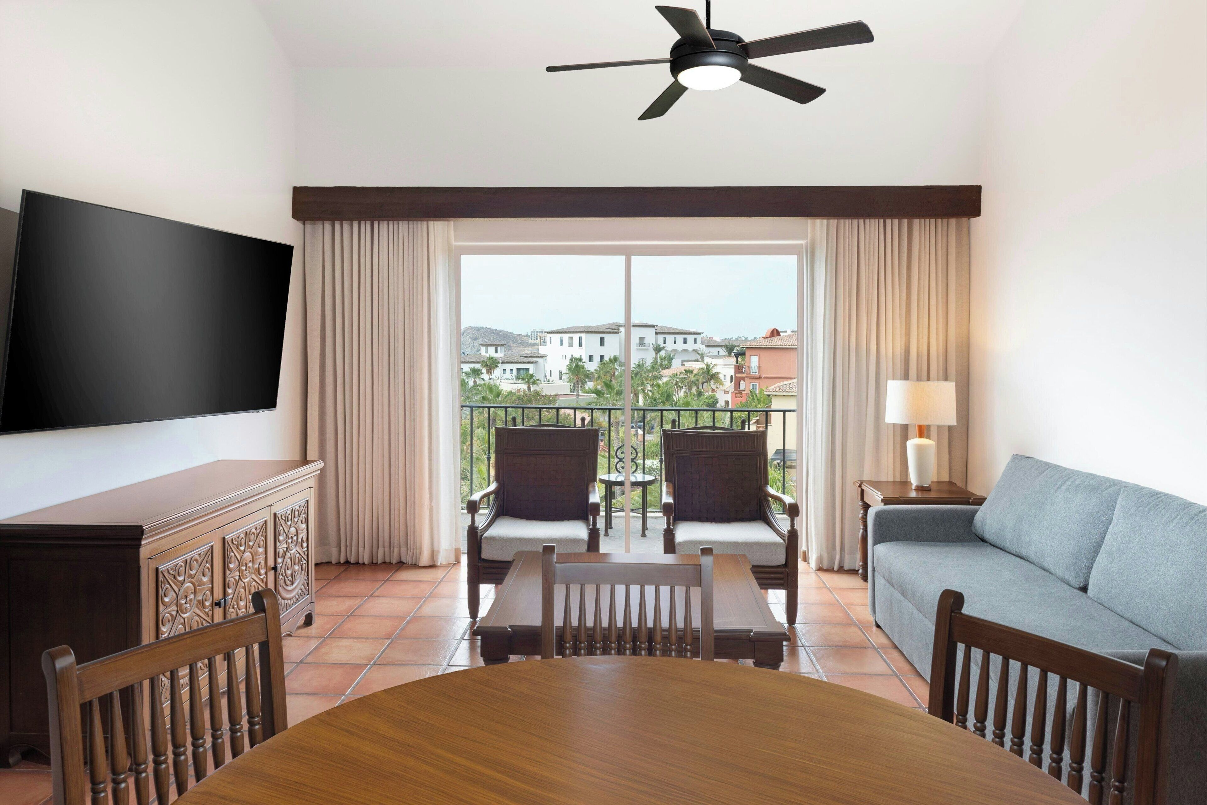 A hotel room with a dining table, chairs, a sofa, a TV, a balcony with chairs, and curtains.