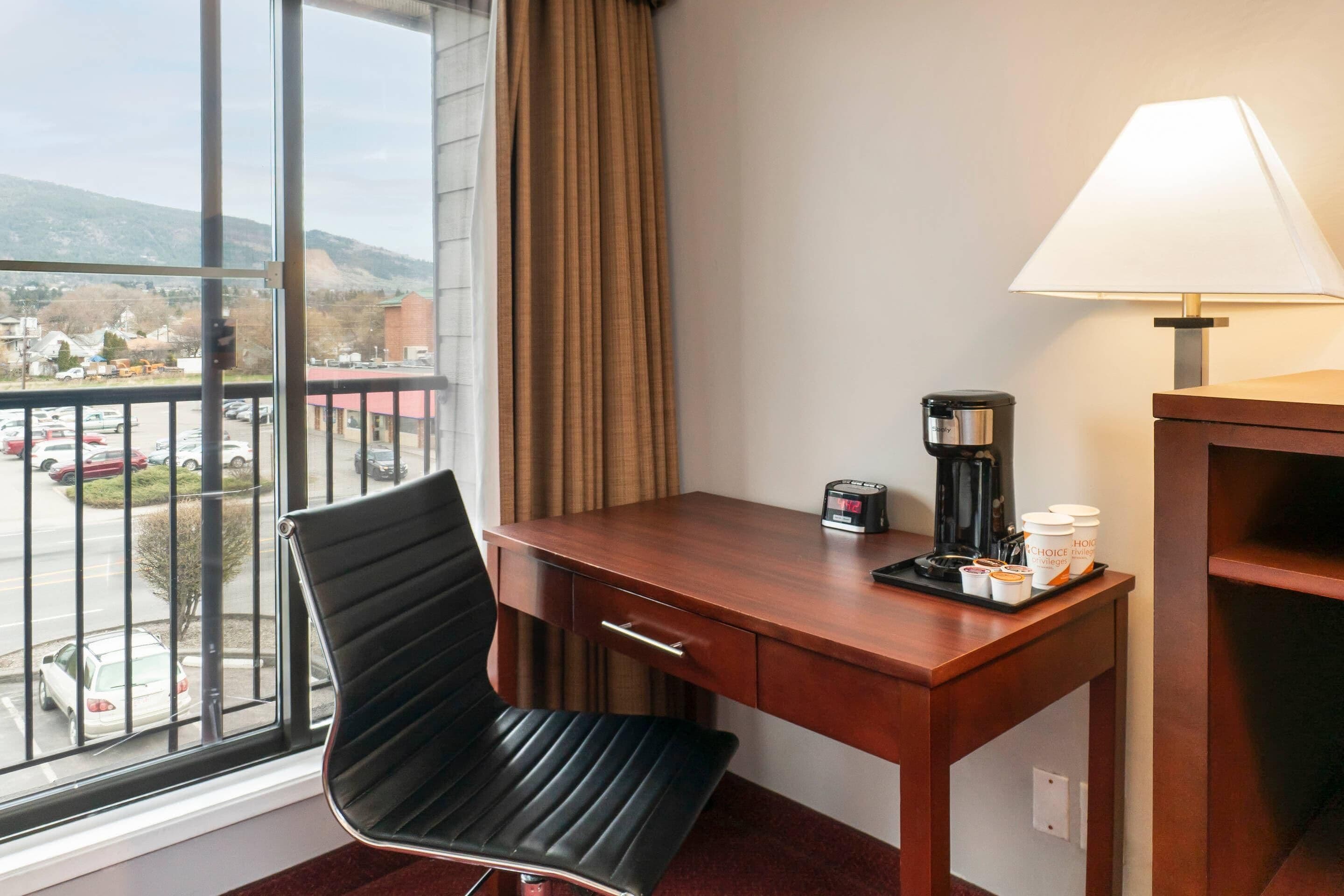 Habitación de hotel con escritorio, silla, lámpara, cafetera y vistas al estacionamiento y a las montañas.