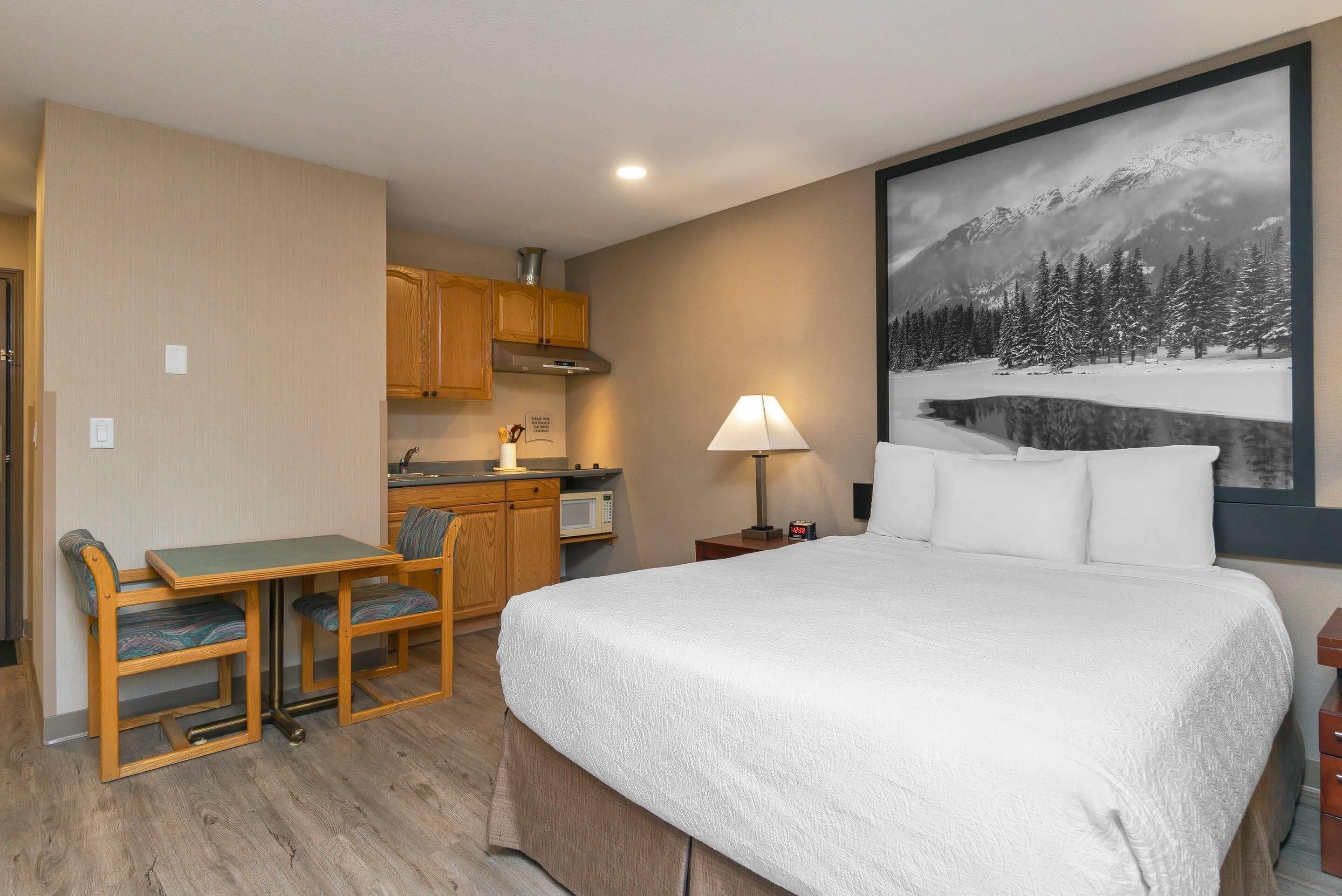 Ein Hotelzimmer mit einem Bett, einem Schreibtisch mit Stühlen, einer Mikrowelle, einer Lampe und einem großen Gemälde mit Bergpanorama.