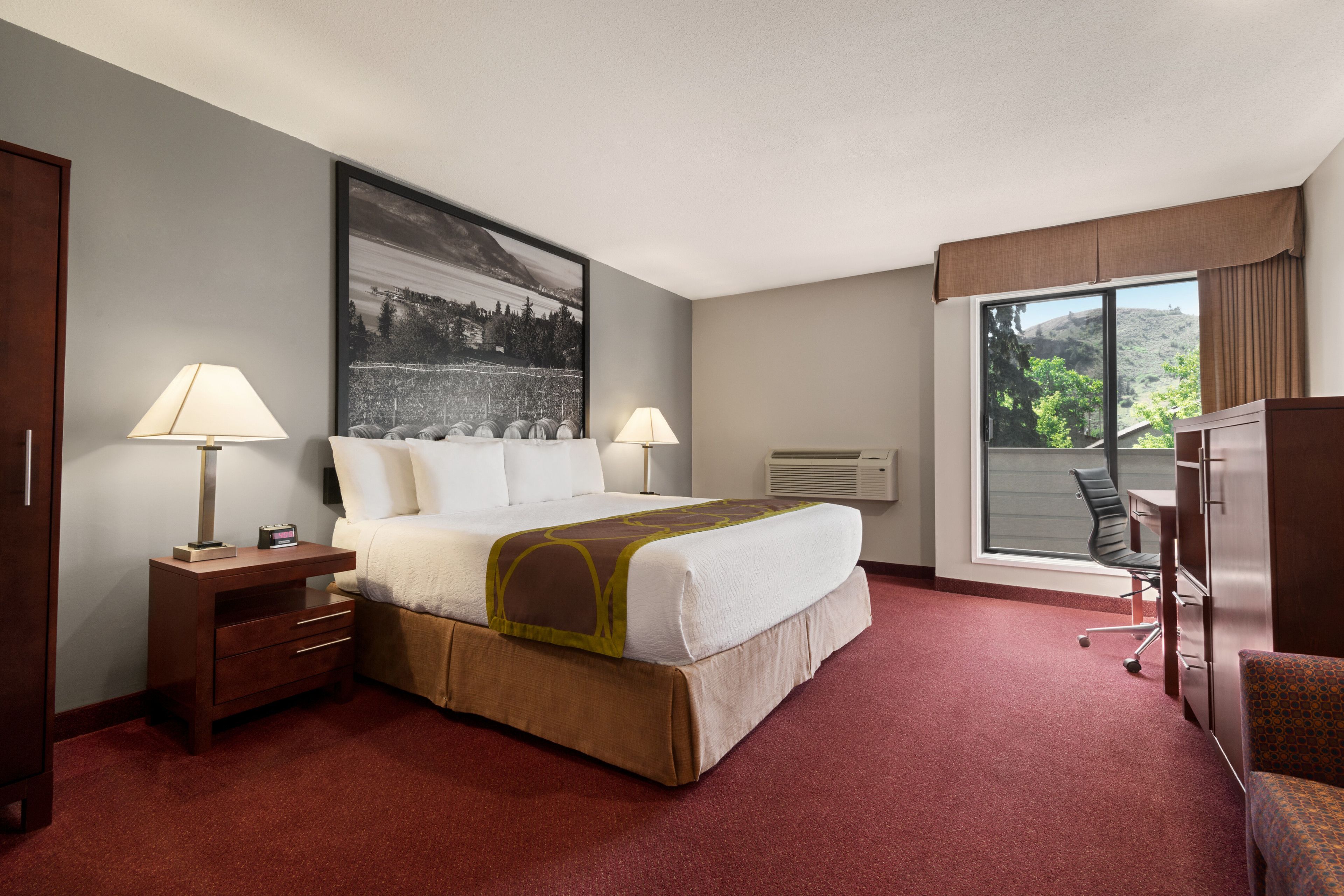 A hotel room with a large bed, two bedside lamps, a desk with a chair, a large window with a view of trees, and a painting on the wall.