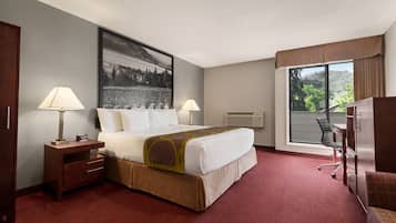 A hotel room with a large bed, two bedside lamps, a desk with a chair, a large window with a view of trees, and a painting on the wall.