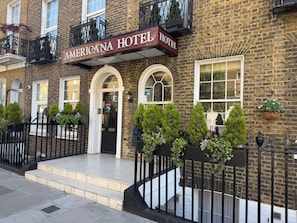 A brick building with a sign reading 'AMERICANA HOTEL'. There are steps leading to the entrance, a black metal fence, and greenery in planters. - Americana Hotel (London)