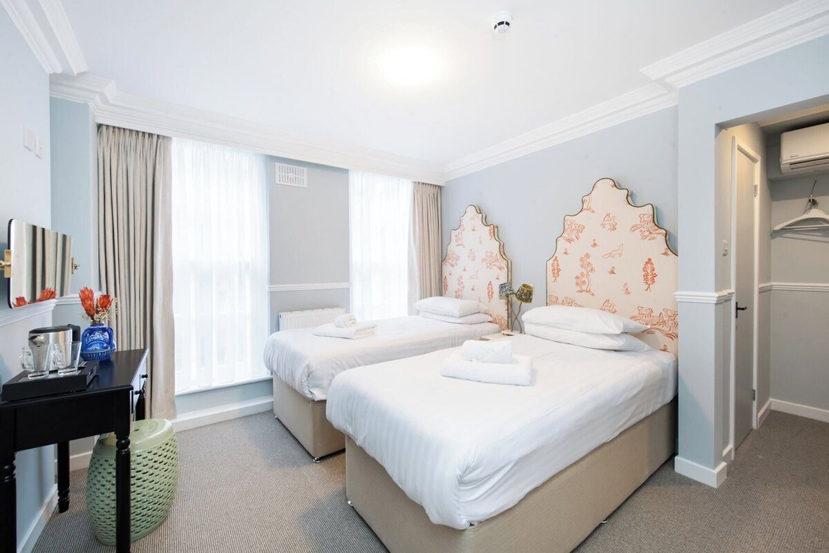 A hotel room with two beds, a desk with a vase, and a radiator.