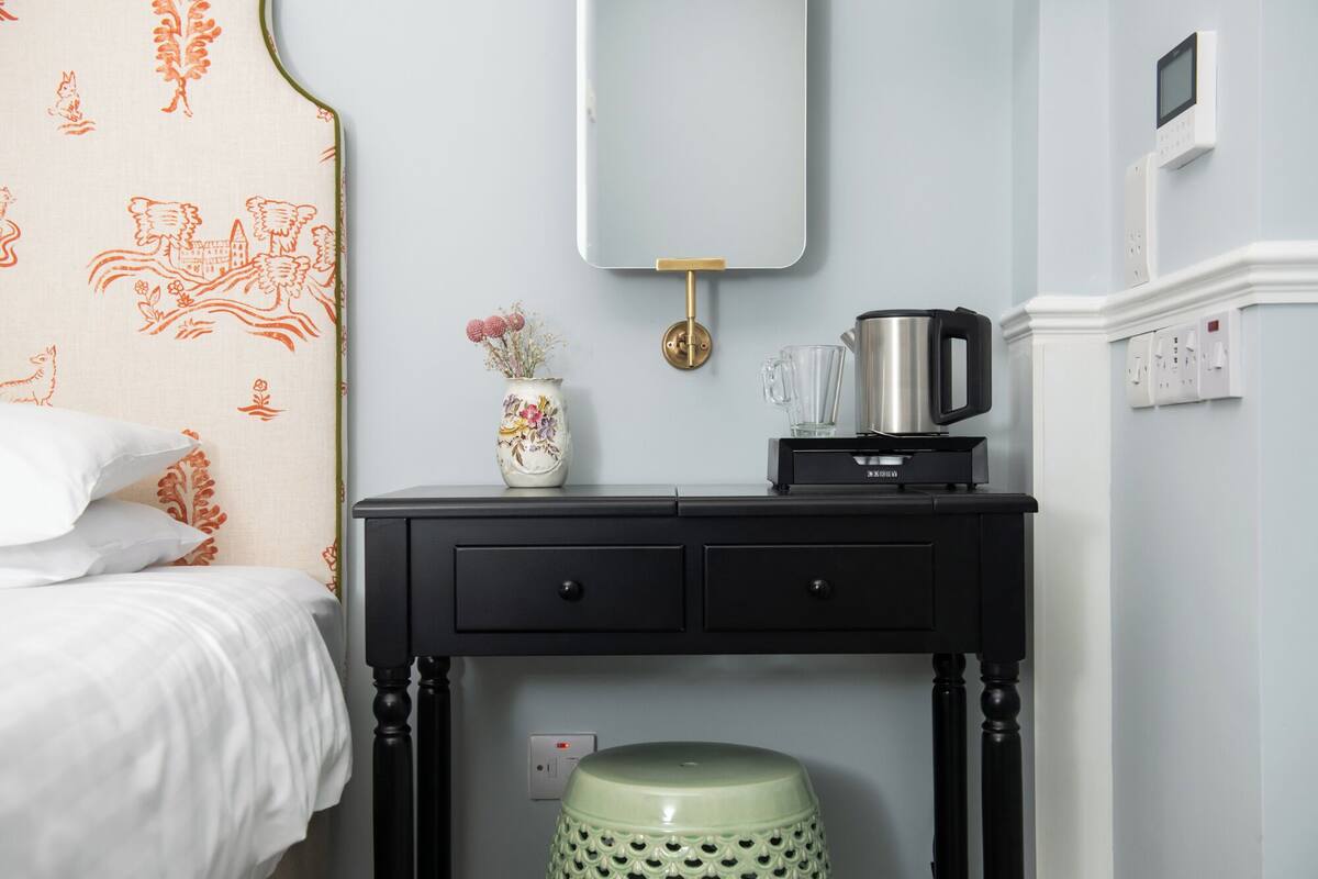 A hotel room with a bed, a small desk with a kettle, a vase with flowers, and a green stool.