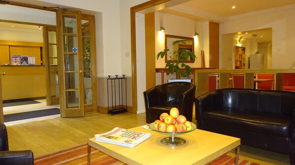 A lobby with a reception desk, a black leather sofa, a glass table with apples, and a newspaper. - Americana Hotel (London)