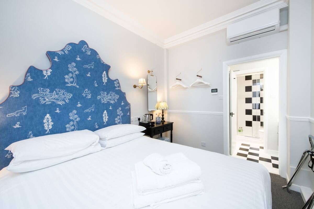 A hotel room with a large bed, a blue headboard with white patterns, a bedside table with a lamp, and a door leading to a bathroom.