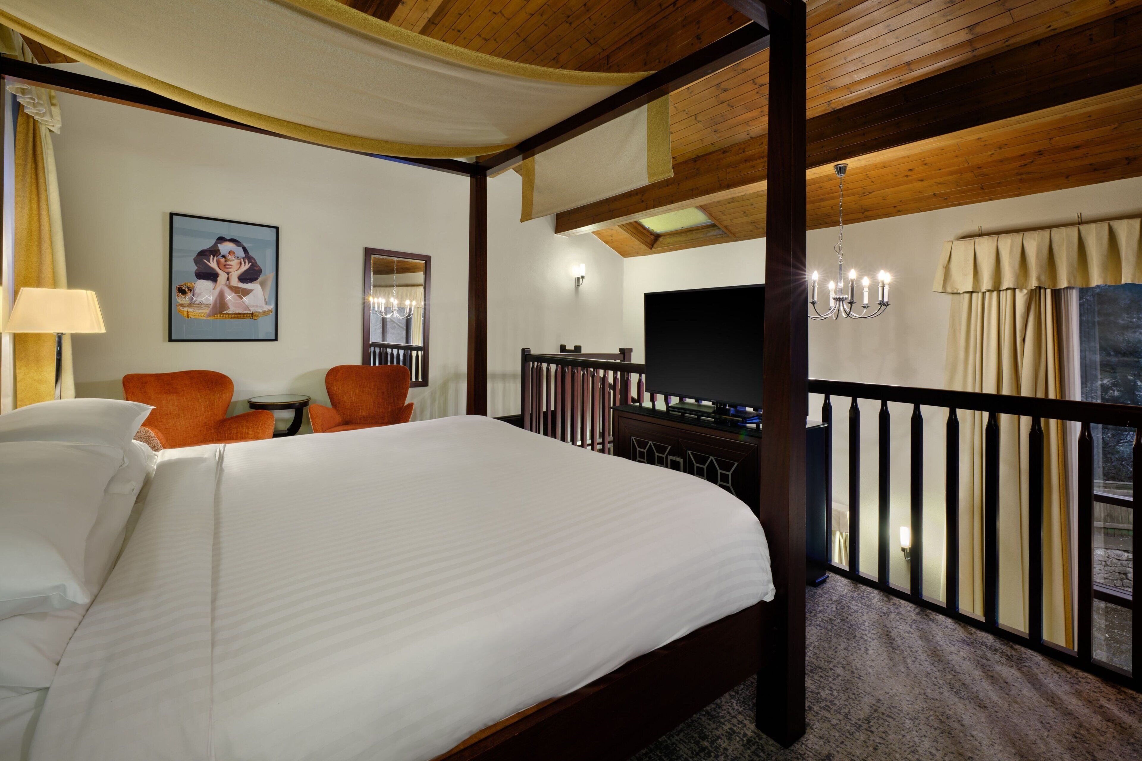 A hotel room with a large bed, a TV, a balcony with a railing, and a chandelier.