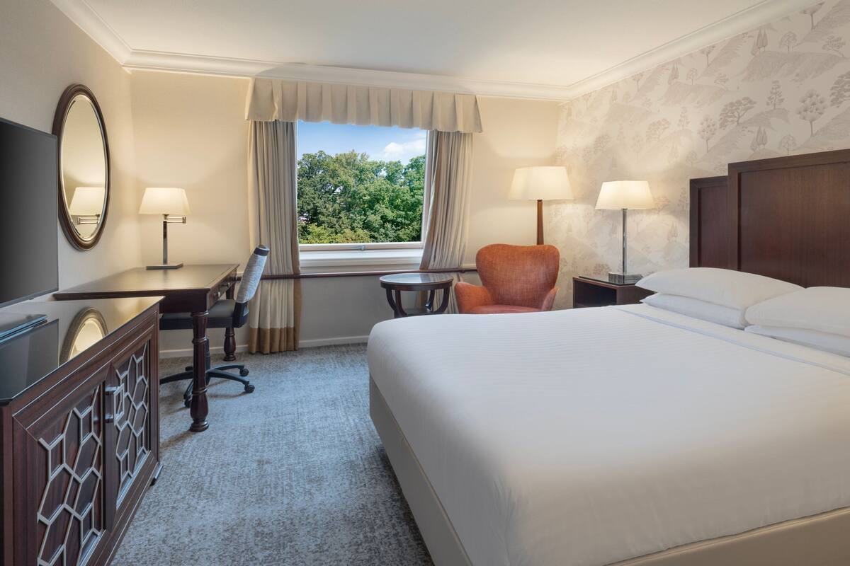 A hotel room with a large bed, a desk with a chair, a small table, two lamps, a mirror, and a window with curtains.