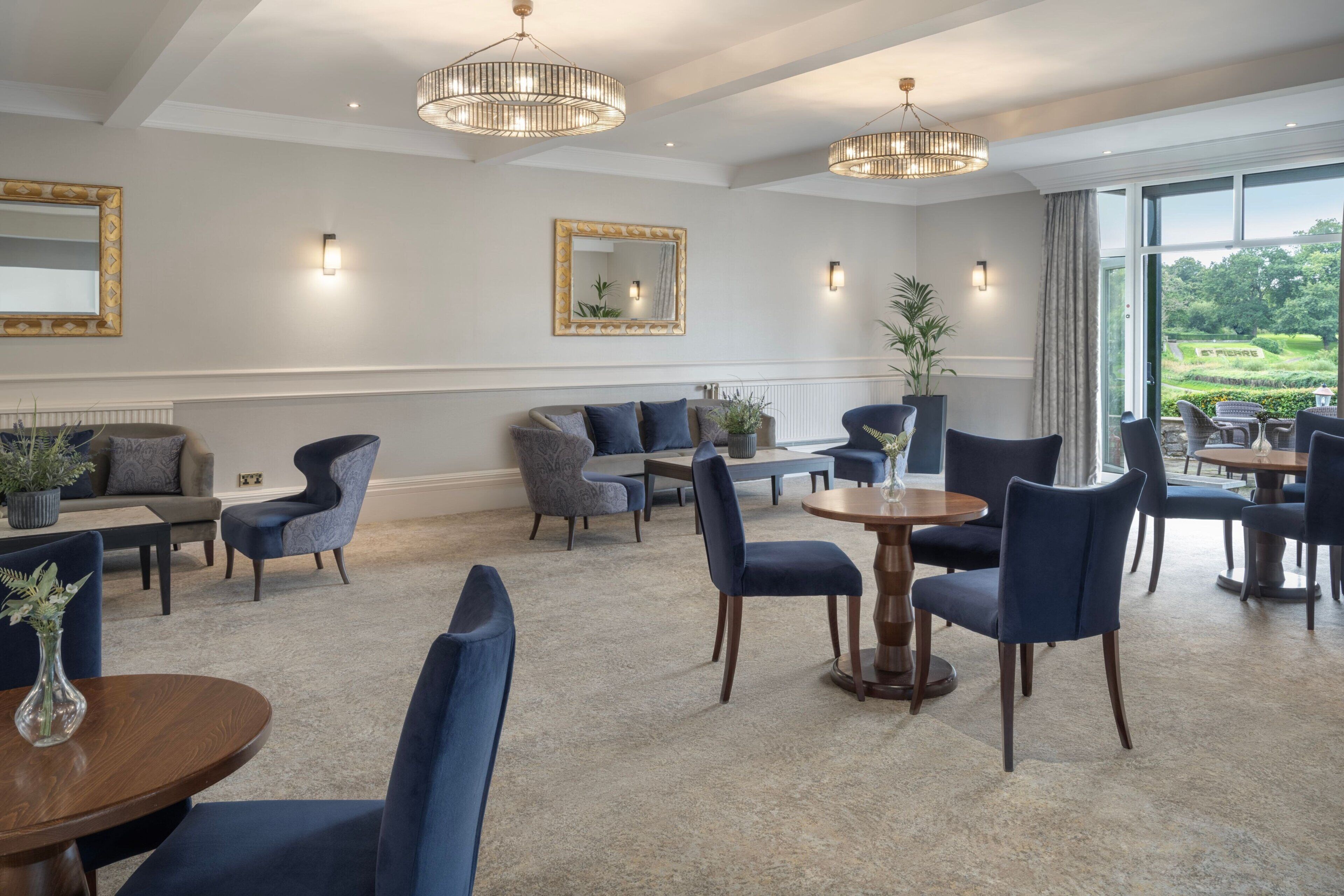 A spacious hotel lobby with a high ceiling, elegant chandeliers, comfortable seating, and large windows offering a view of the outdoors.
