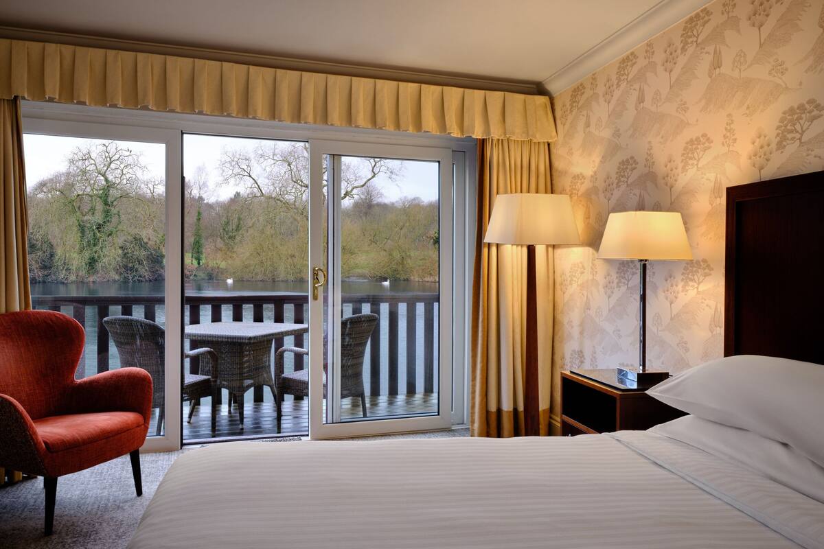 A hotel room with a bed, a chair, a table, and a balcony with a view of trees and a body of water.