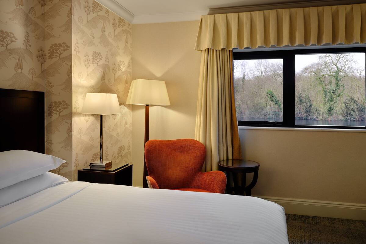 A hotel room with a large bed, two bedside lamps, a red armchair, a small table, and a window with curtains.