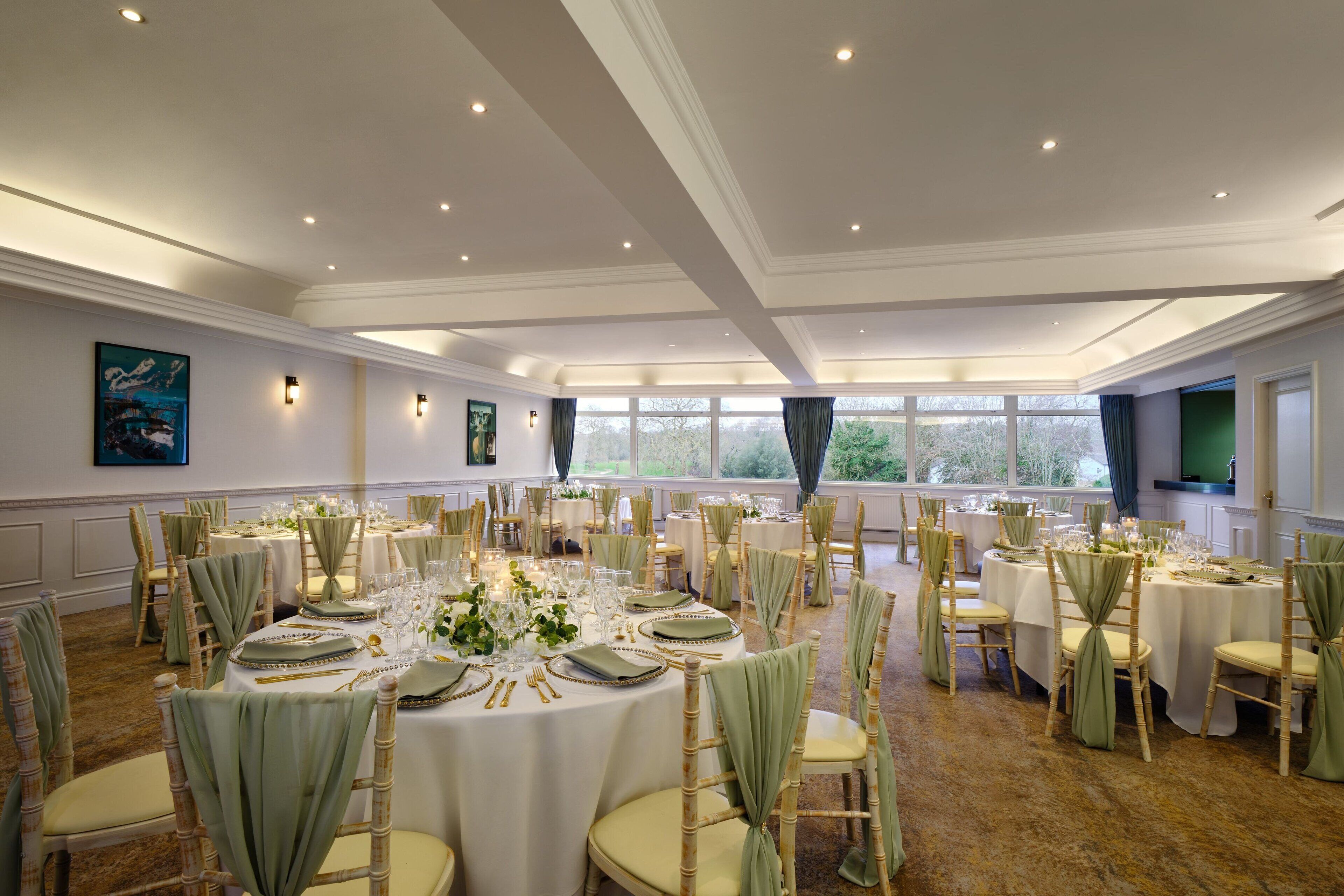 A banquet hall with round tables set for dining, surrounded by chairs with green sashes, and large windows providing natural light.