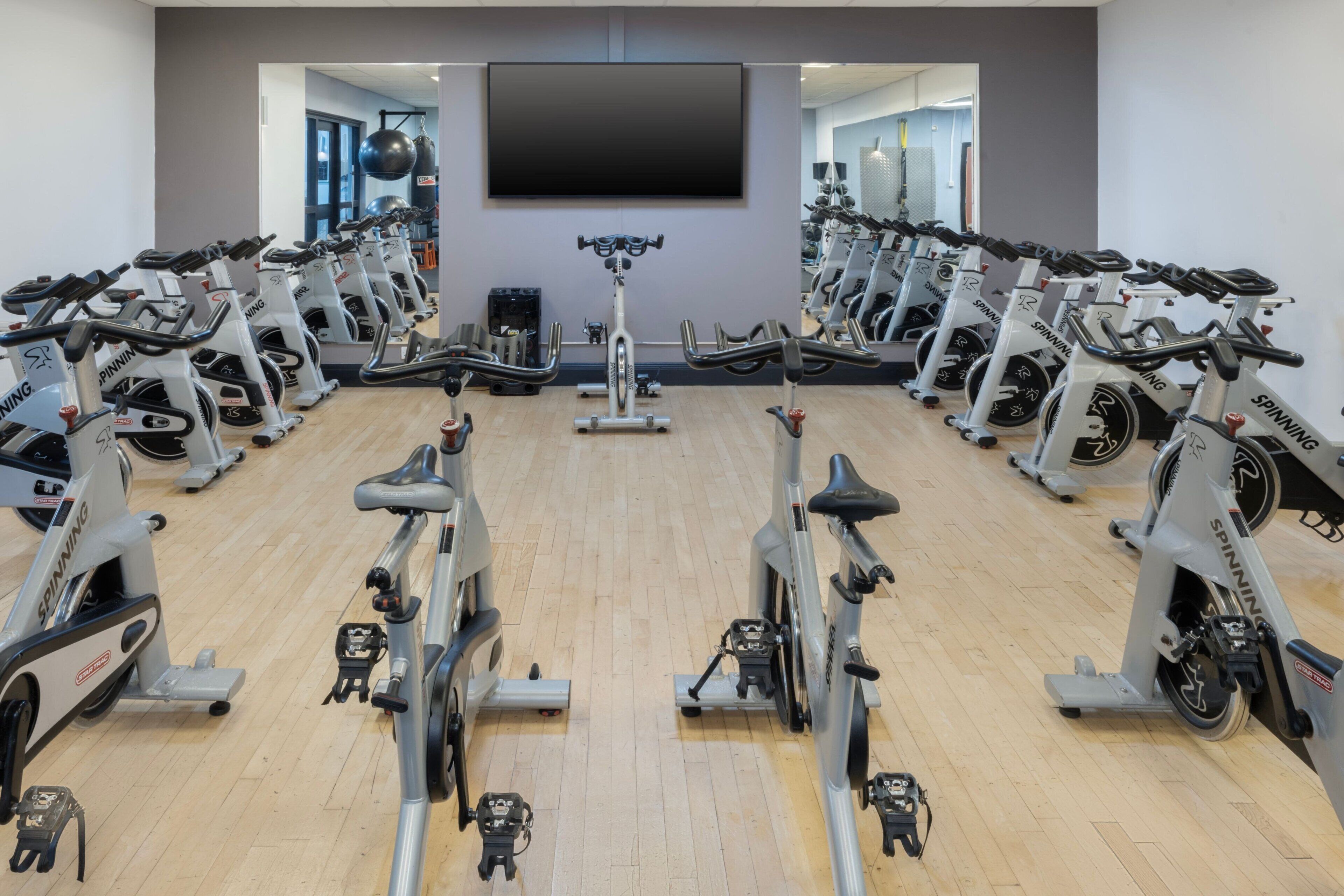 Une salle de sport avec plusieurs vélos d’appartement disposés en rangées, un grand écran de télévision au mur et un plancher en bois.