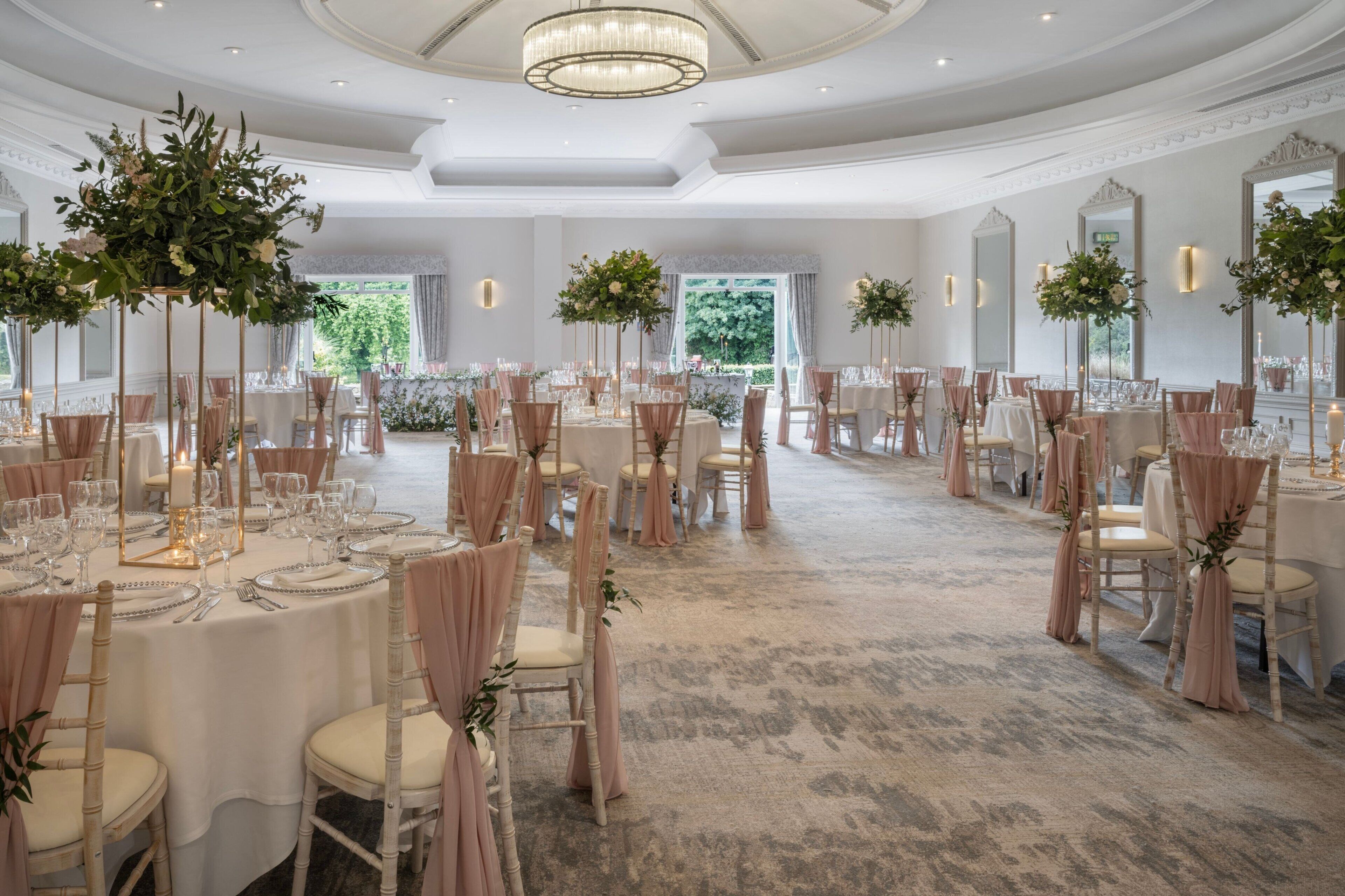 A spacious banquet hall with round tables, elegant chandeliers, and floral centerpieces.