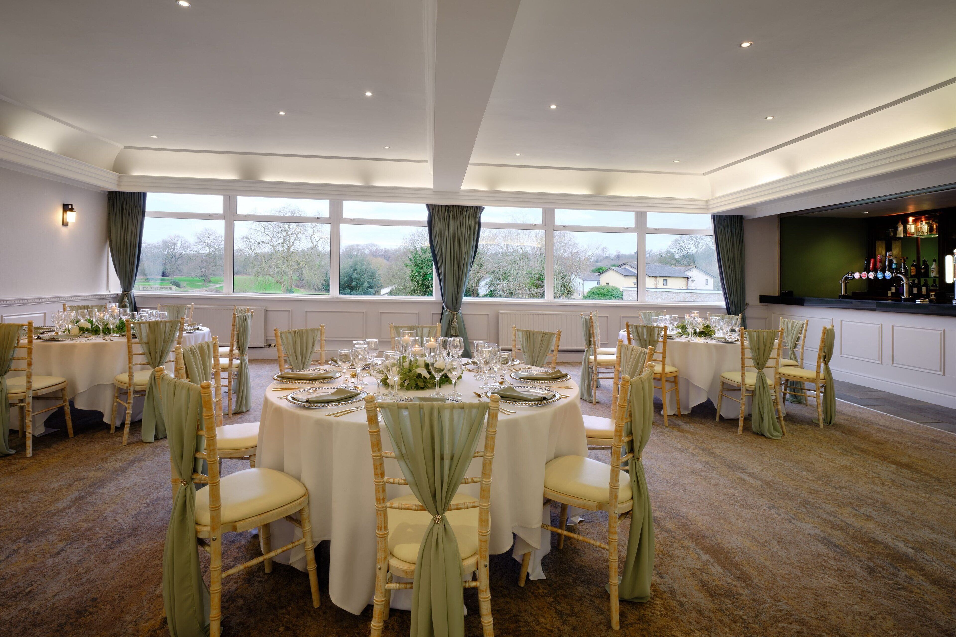 A formal dining room with round tables set for a meal, large windows with curtains, and a bar area with bottles.