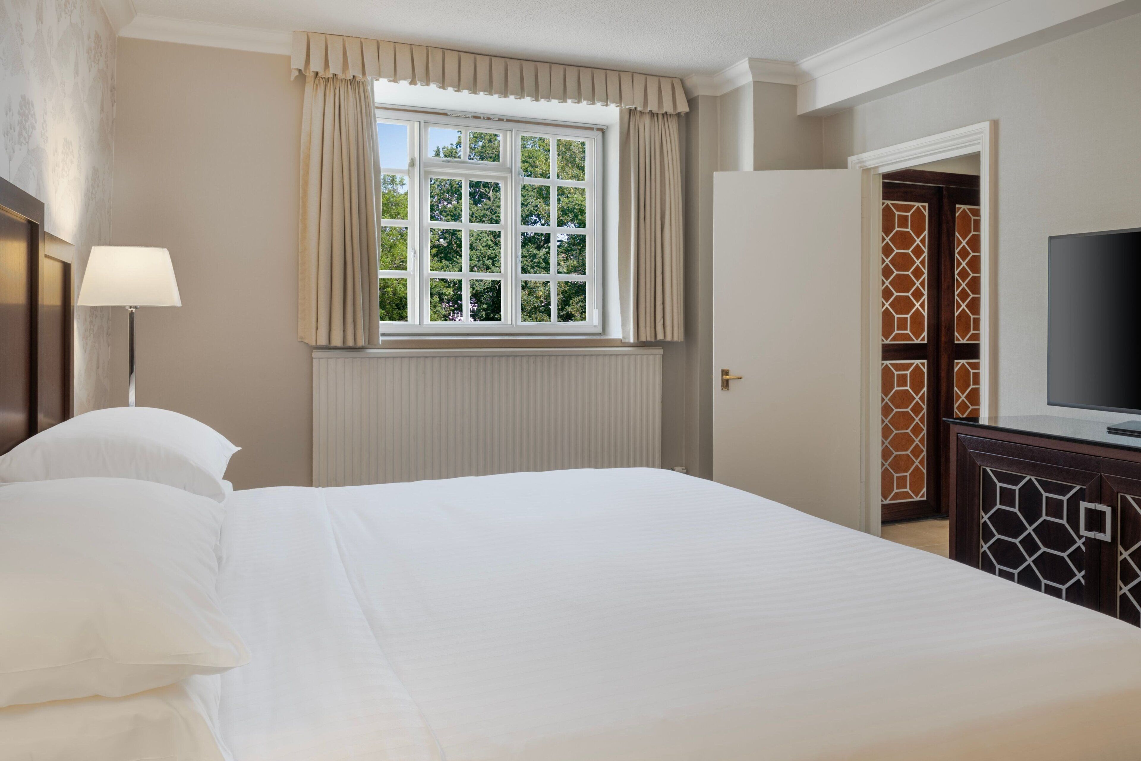 A hotel room with a large bed, a window with curtains, a bedside lamp, a television, and a wardrobe with a geometric pattern.