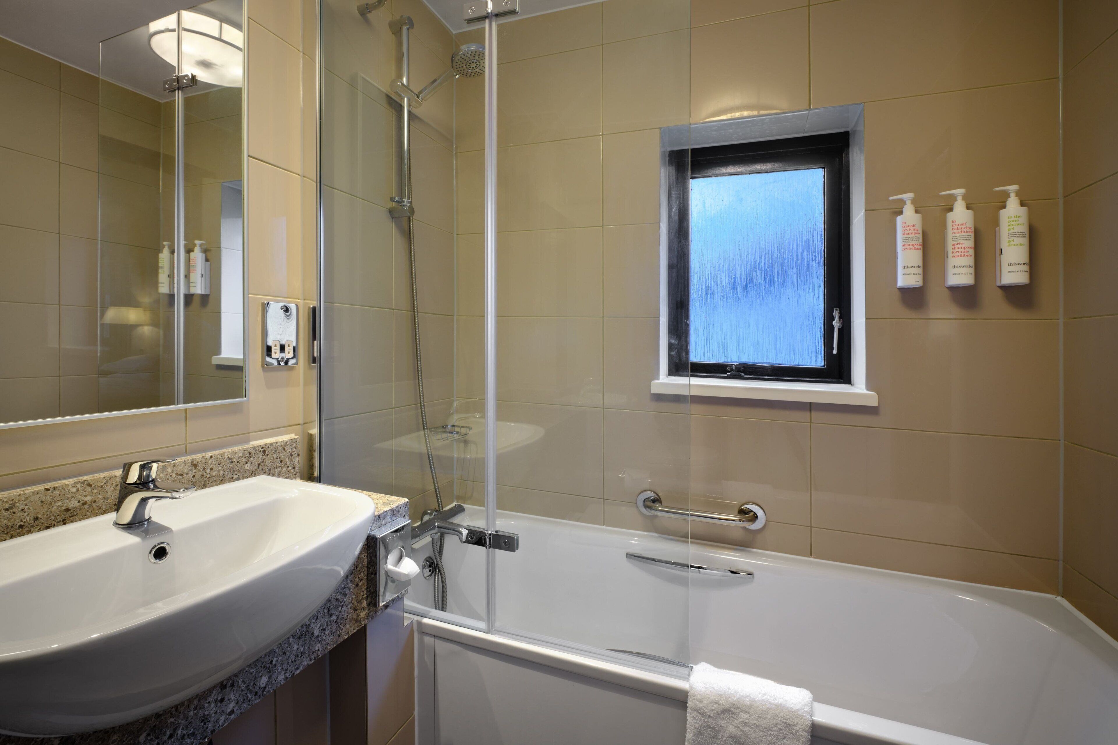 Ein Badezimmer mit Waschbecken, einer Duschkabine, einem Fenster und Pflegeprodukten an der Wand.