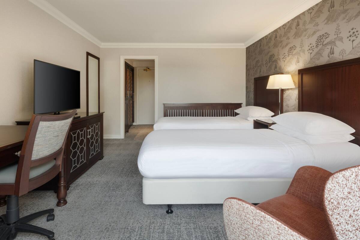 A hotel room with two beds, a desk with a TV, a chair, and a lamp.