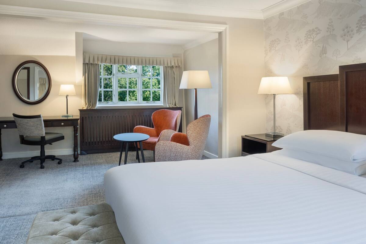 A hotel room with a bed, two chairs, a desk, a lamp, and a window with curtains.