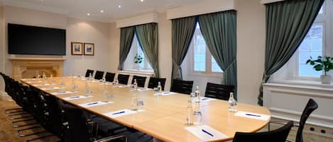 Un salón de conferencias con una larga mesa, sillas, un televisor de pantalla plana y ventanas con cortinas.