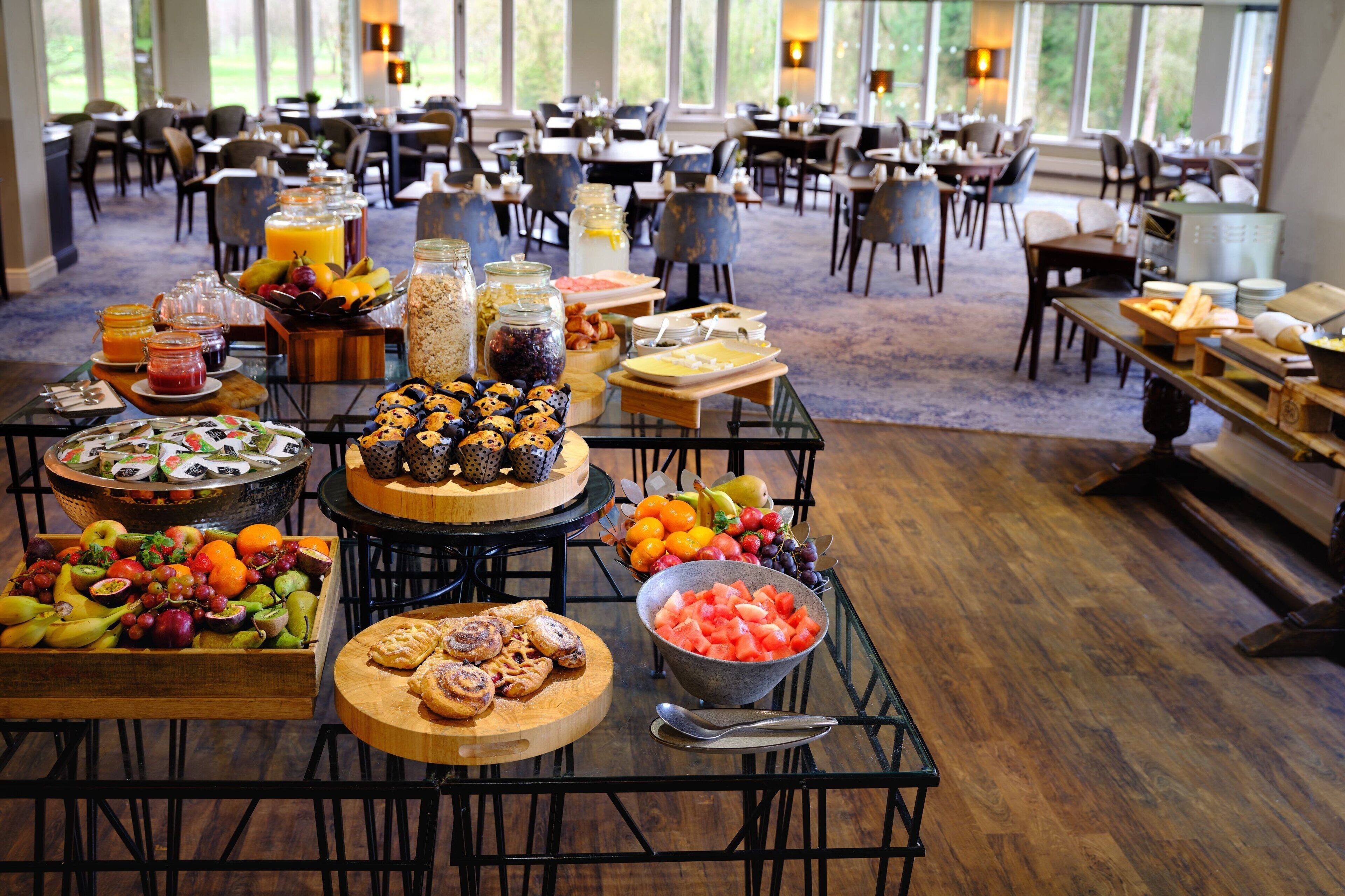 A buffet with various fruits, pastries, and beverages spread across multiple tables in a spacious dining area with large windows.