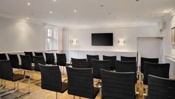 A conference room with rows of black chairs, a flat-screen TV mounted on the wall, and a window on the left side.