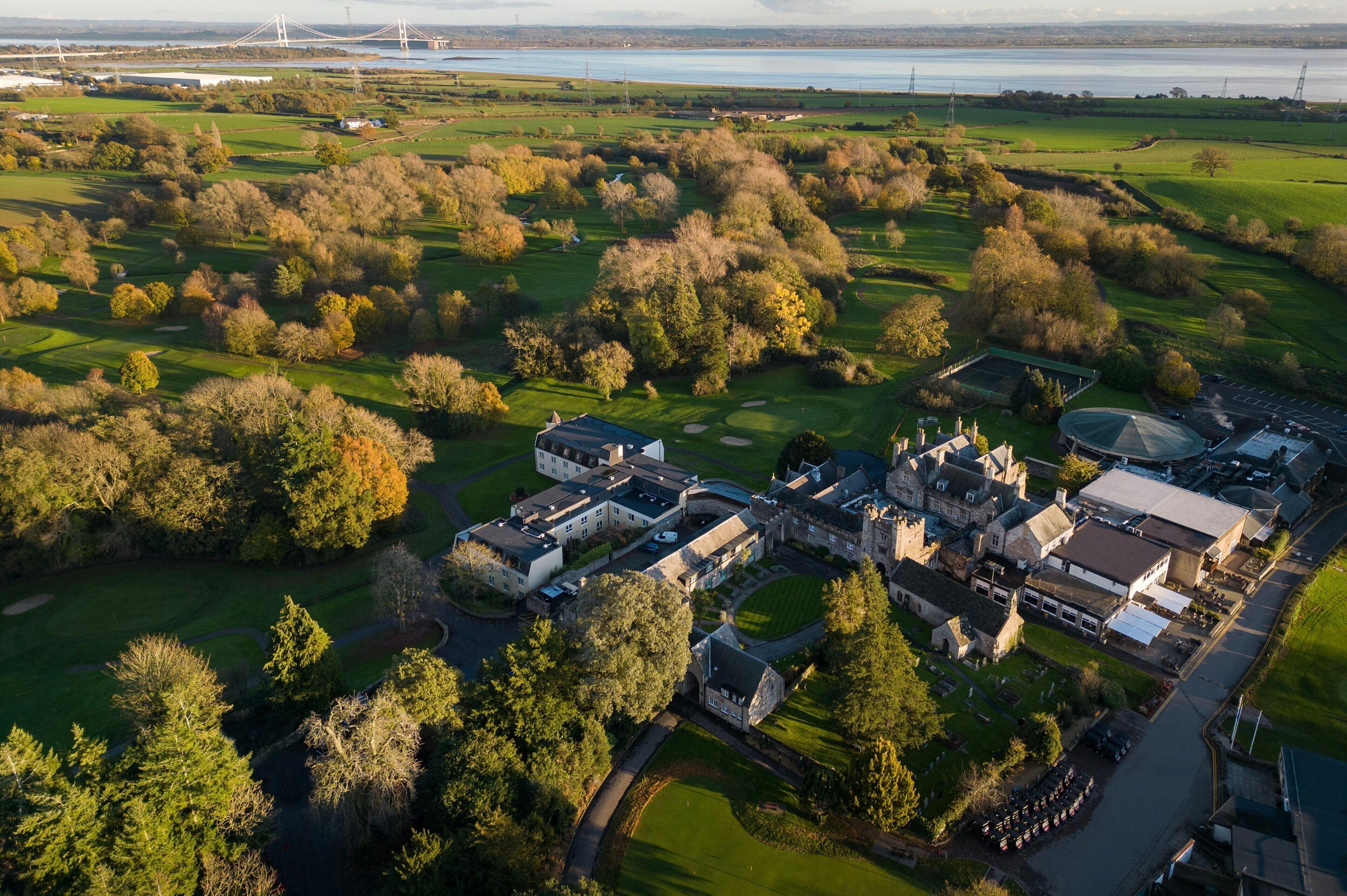 An aerial view of a golf course with a clubhouse, a parking area, and a large body of water in the background.