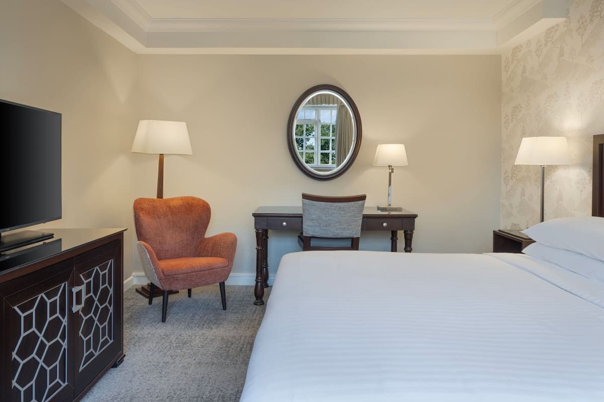 A hotel room with a large bed, a desk with a chair, a television on a cabinet, and two lamps.