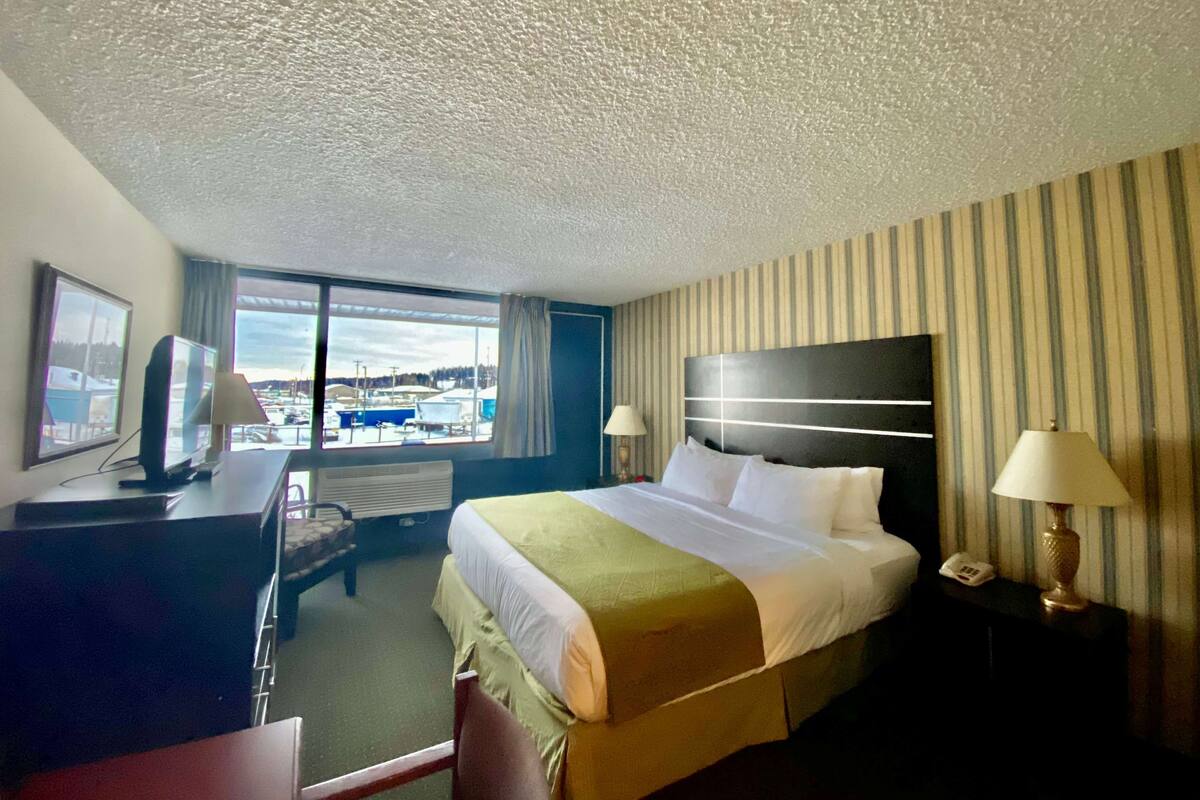 Ein Hotelzimmer mit einem großen Bett, einem Schreibtisch mit Computer, einem Telefon und einem Fenster mit Blick nach draußen.