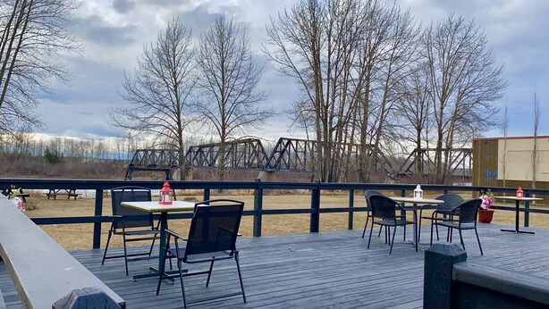 An outdoor deck with tables and chairs, overlooking a bridge and trees. - Ramada by Wyndham Whitecourt (Whitecourt)