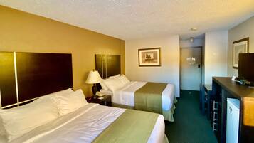 A hotel room with two beds, a desk, a chair, a lamp, and a refrigerator.