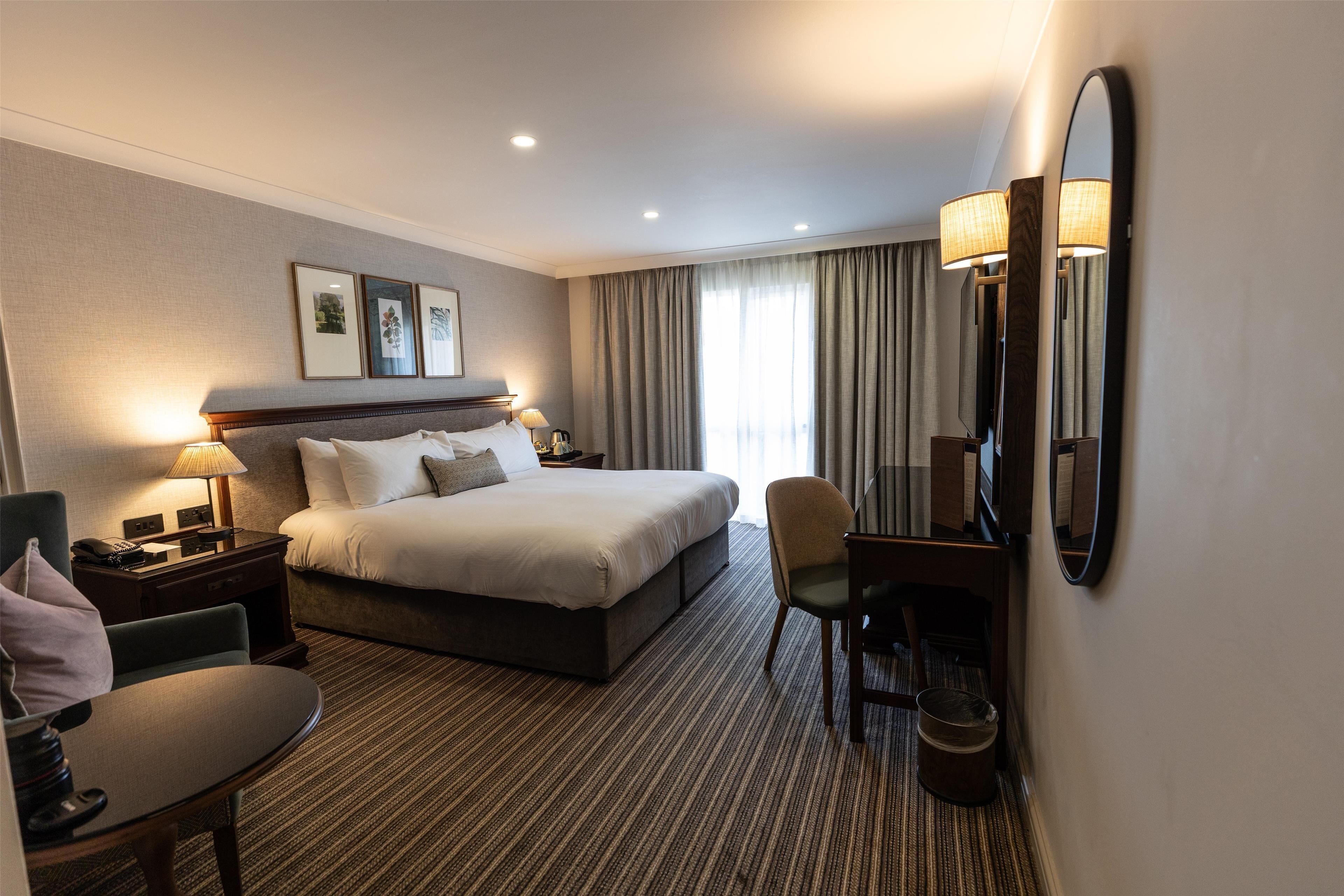 A hotel room with a large bed, bedside tables with lamps, a desk with a chair, and a large mirror.