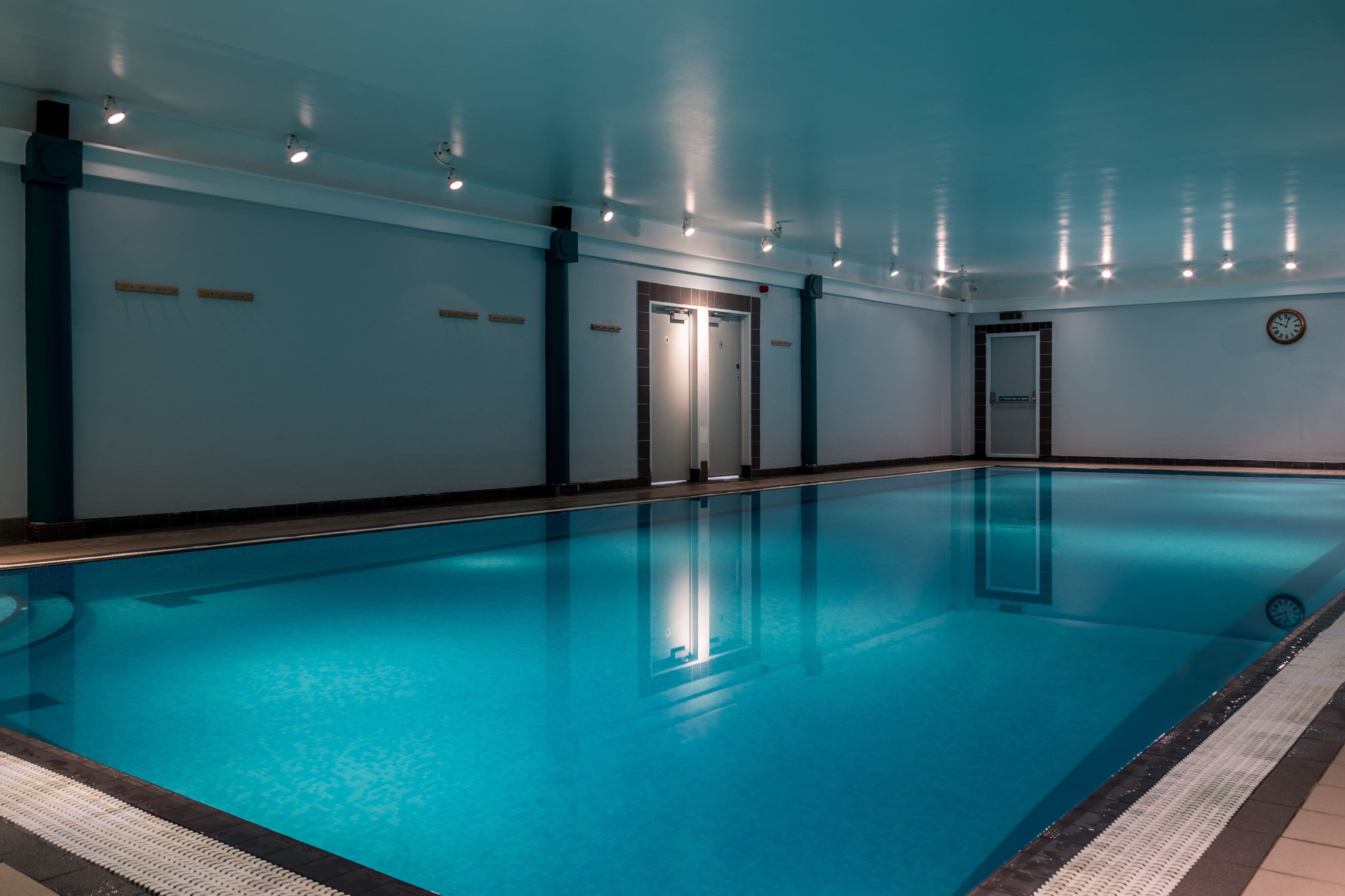 Une grande piscine intérieure à l’eau claire, entourée de murs sur lesquels sont affichées une horloge et des panneaux indiquant les sorties de secours.
