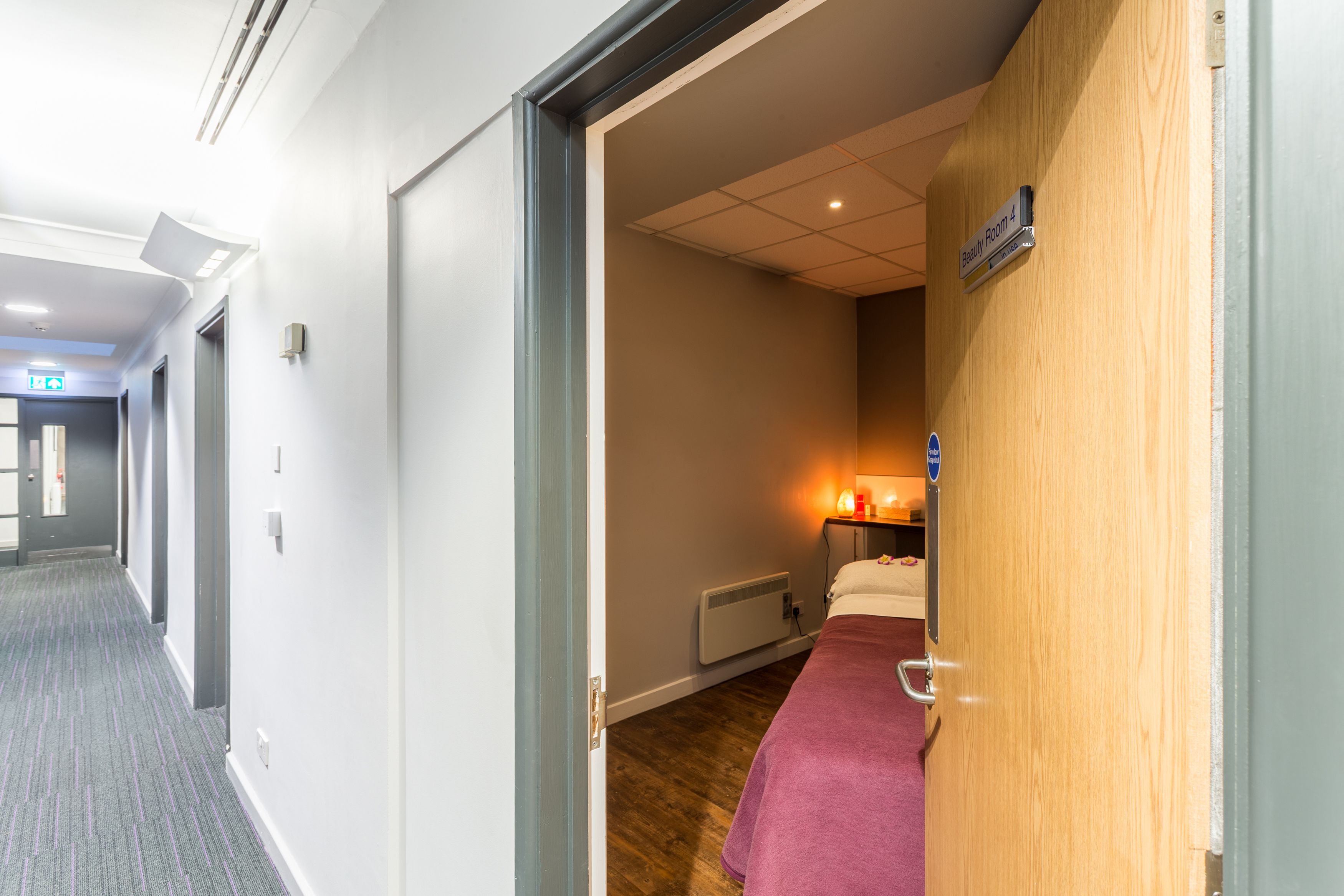 A hotel room with a wooden wall, a bed with a pink blanket, a bedside table with a lamp, and a sign indicating the room number.