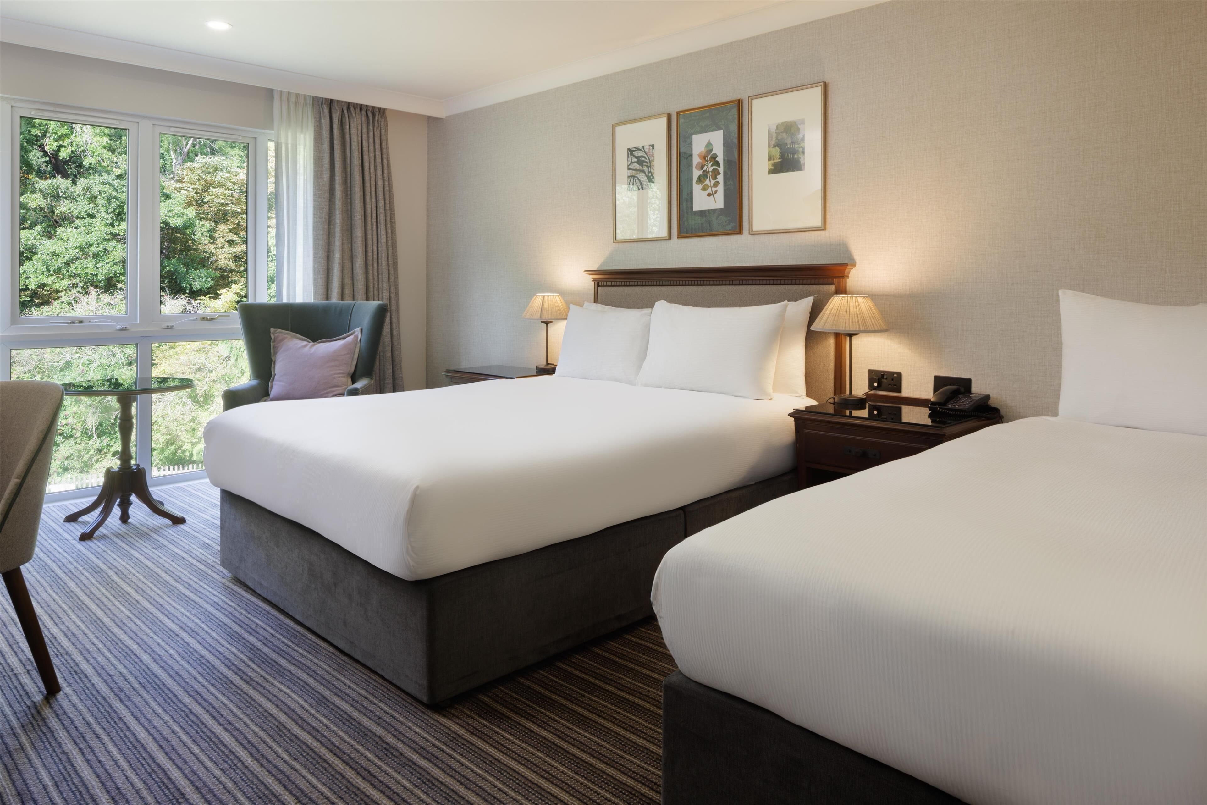 A hotel room with two beds, a chair, a small table, and a window with a view of greenery.