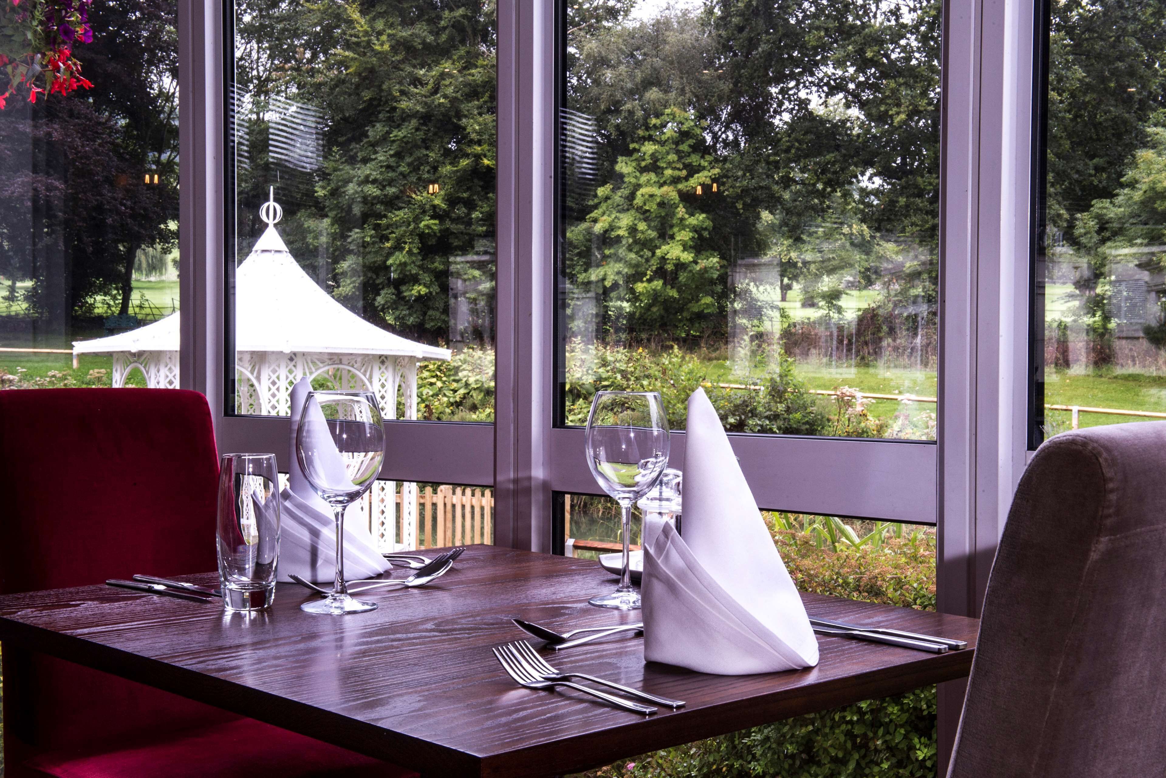 A dining table set with cutlery, glasses, and a napkin, with a view of a garden and a gazebo through large windows.