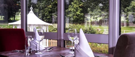 A dining table set with cutlery, glasses, and a napkin, with a view of a garden and a gazebo through large windows.
