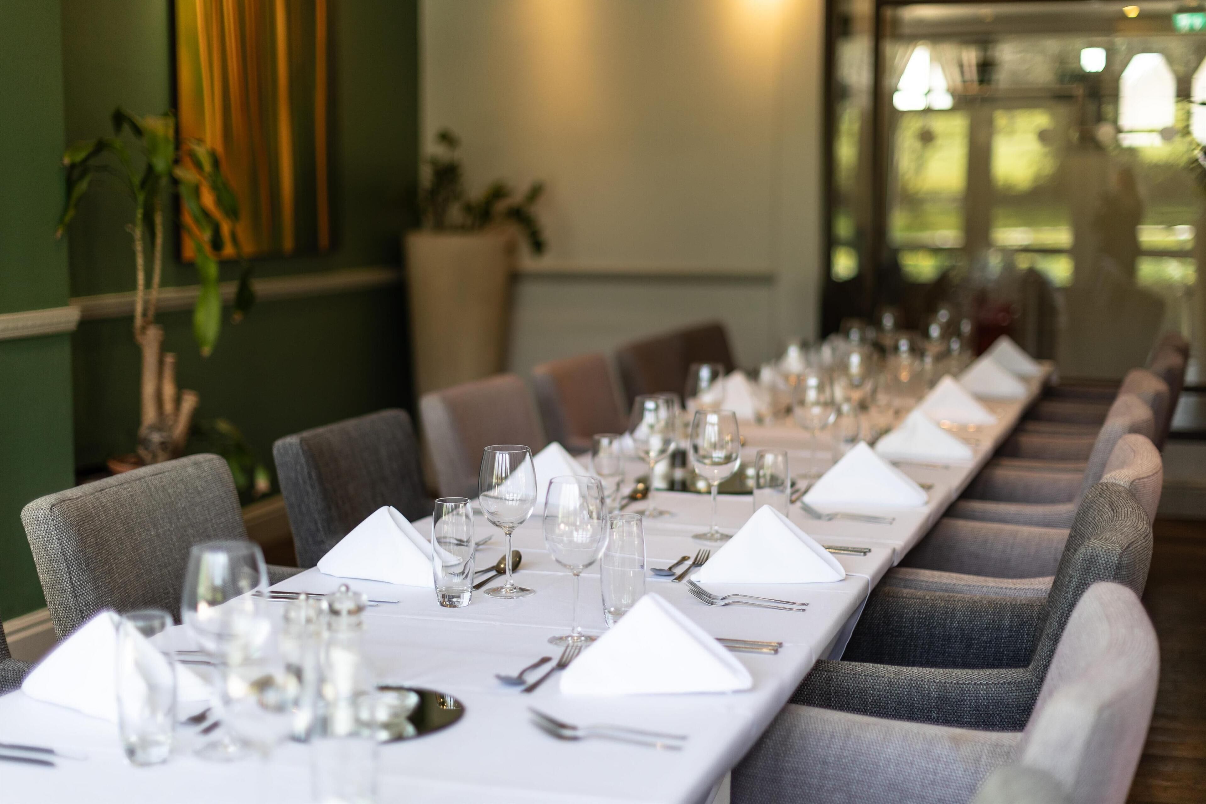 A long dining table set with white tablecloths, silverware, and wine glasses, flanked by upholstered chairs.